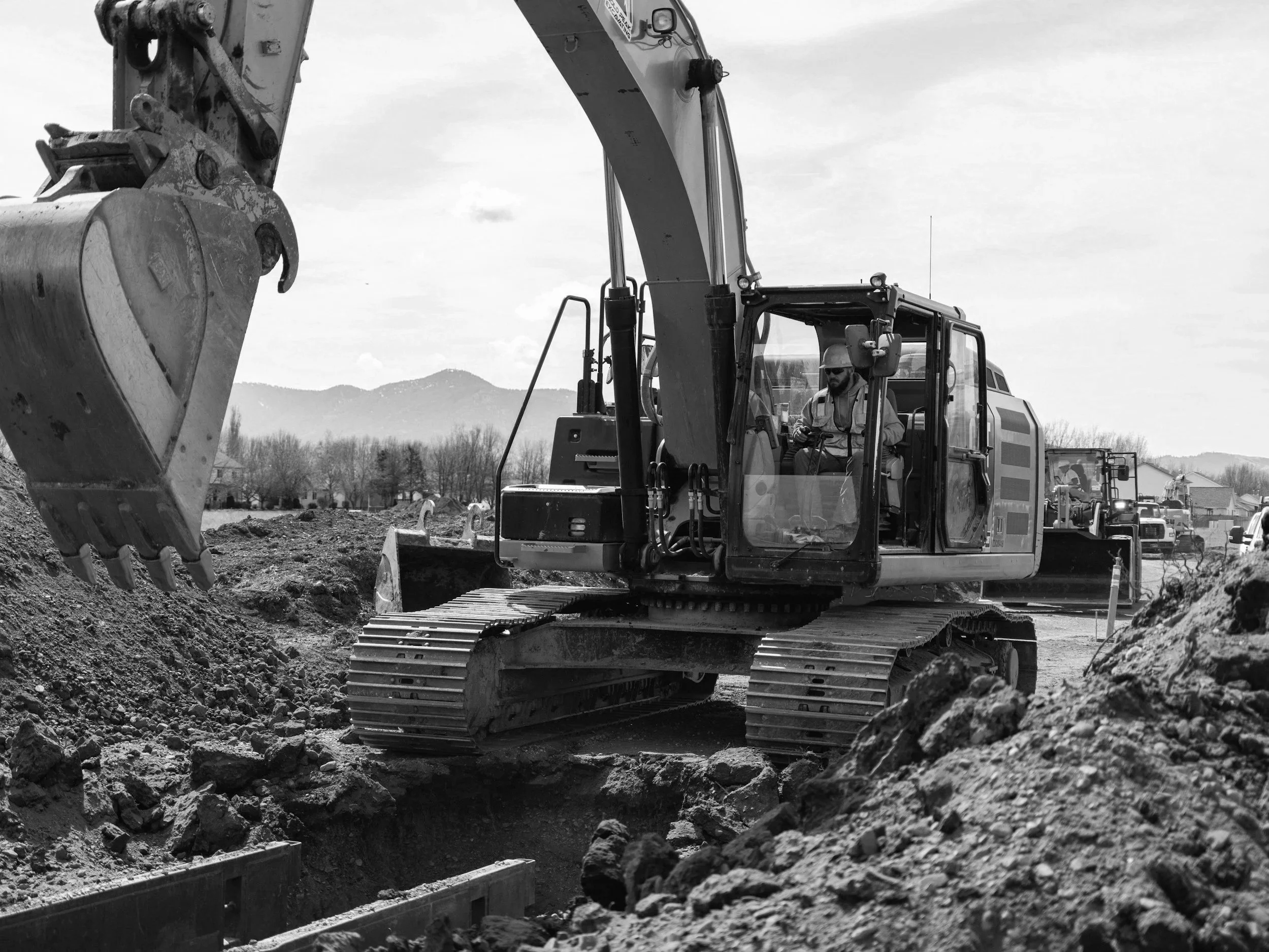 Black and white photo of an excavator digging in a construction site with mountains in the background.
