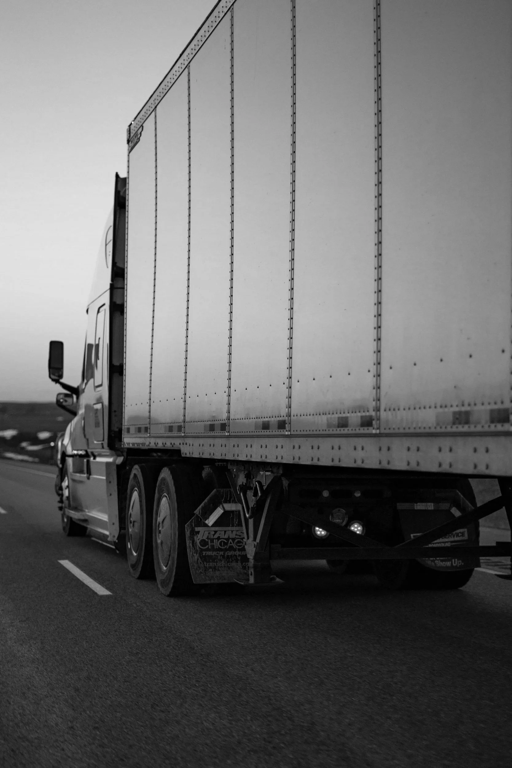 Black and white photo of a semi-truck driving on a highway, capturing the front and side of the truck with visible wheels and cargo trailer.