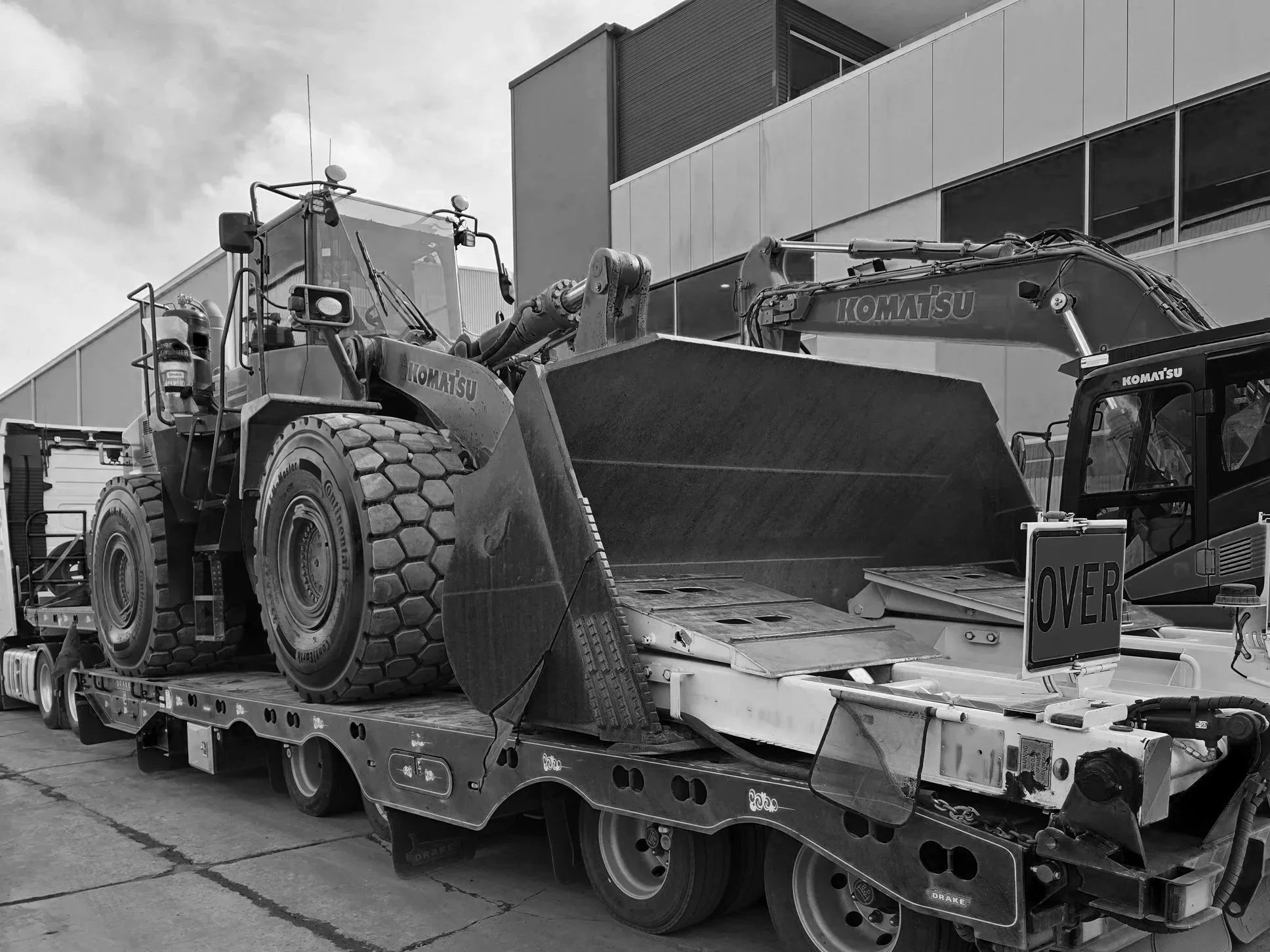 A large Komatsu construction vehicle with a scoop attachment on a flatbed truck, with a modern building in the background.