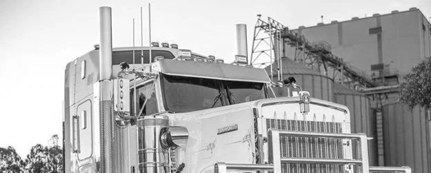Black and white photo of a large semi-truck on the road, with industrial buildings in the background.