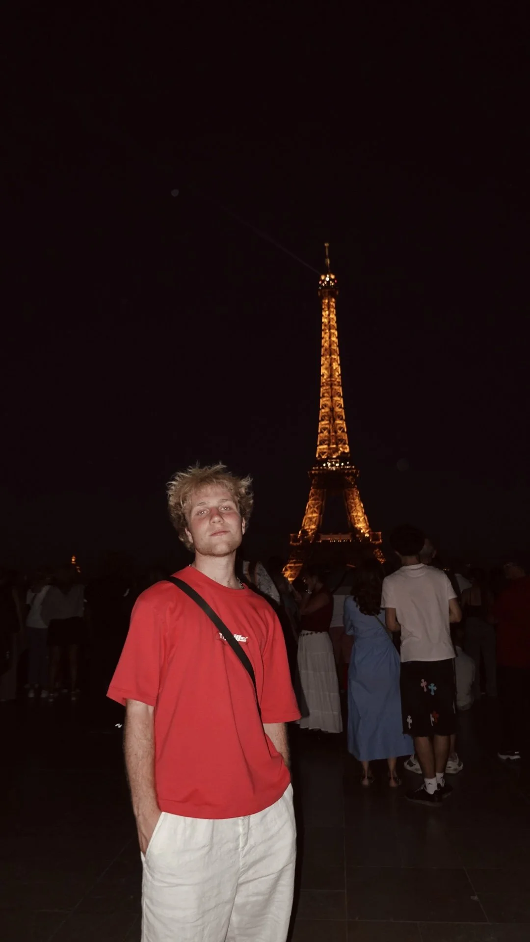 A young man with curly blond hair and a red shirt stands in front of the illuminated Eiffel Tower at night, surrounded by other people.