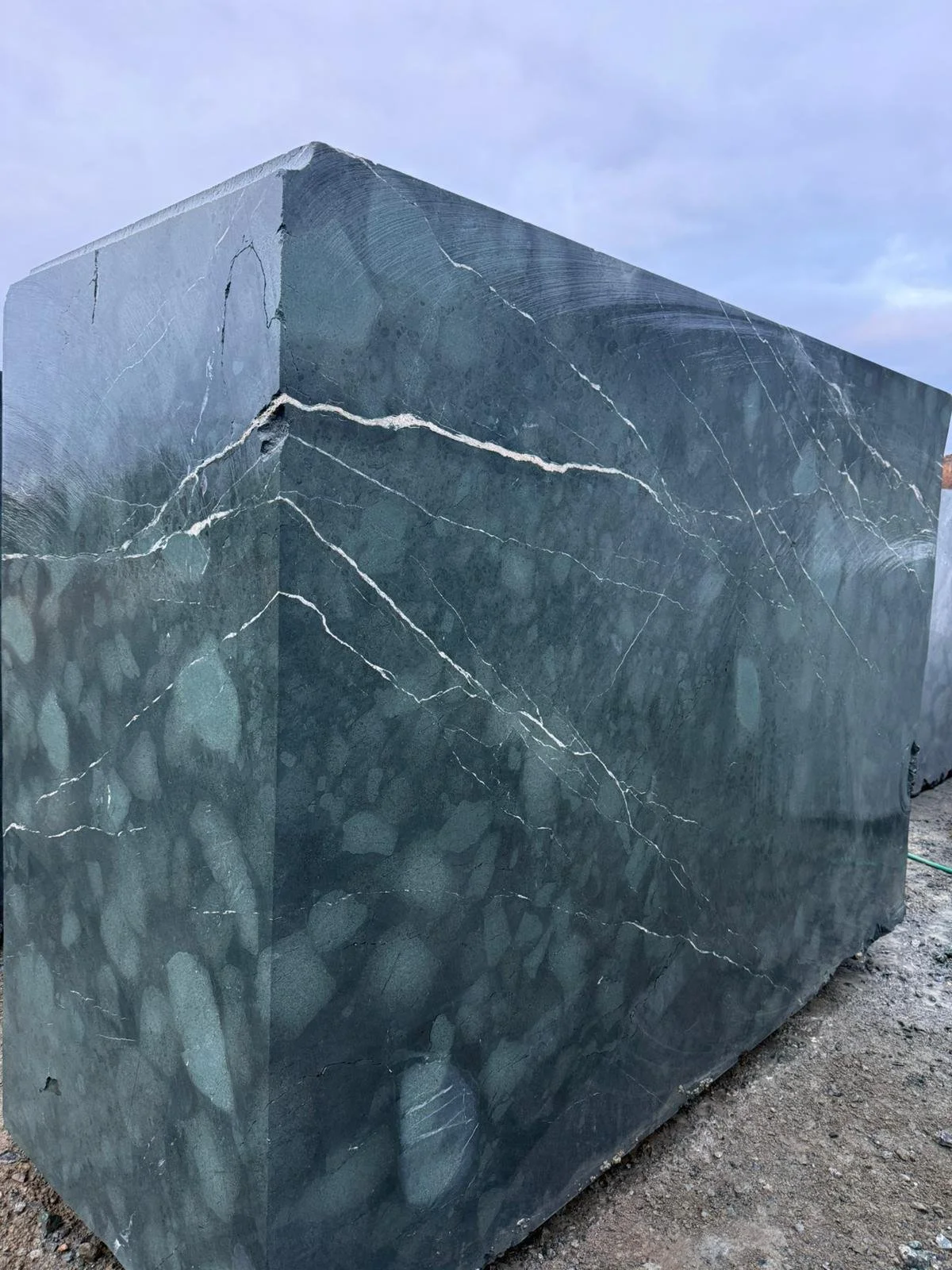 Large polished green marble block with white veining, outdoors under cloudy sky.