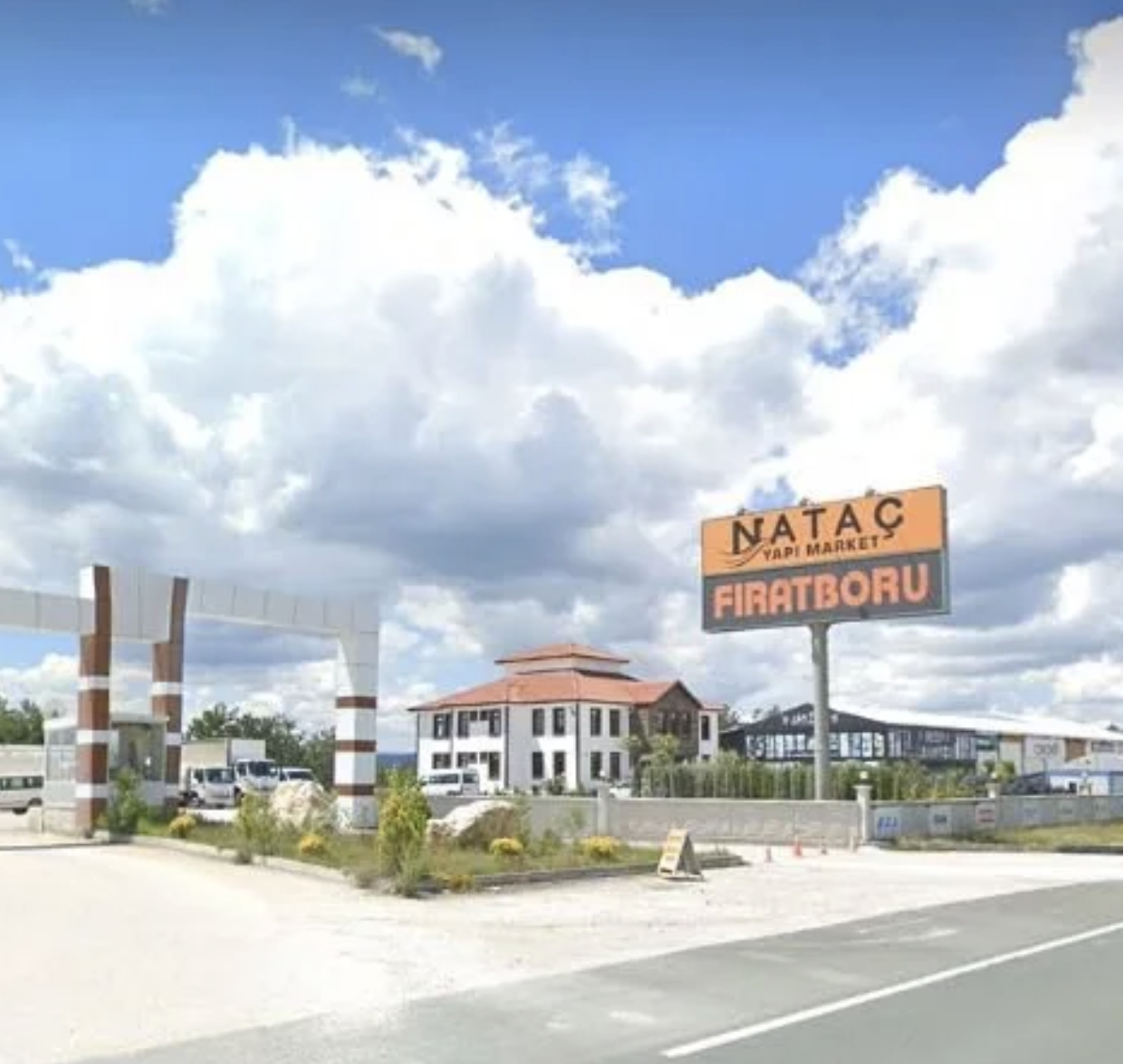A roadside scene with a sign for Nataç Yapı Market and Firatboru, a building with a red roof, and a partly cloudy sky.
