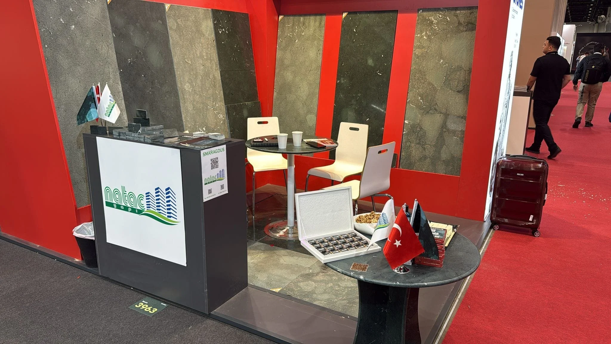 Exhibition booth with a round table, two chairs, flags, a box of snacks, and a display with logos, set on a red and gray booth at an event.
