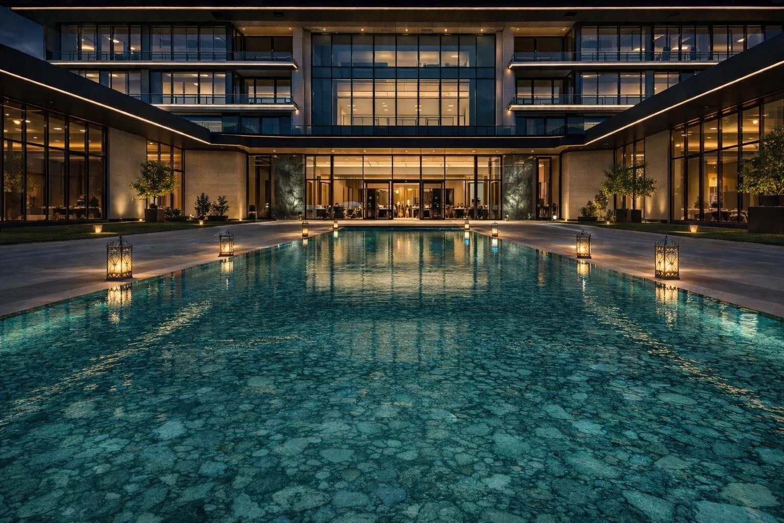 Luxurious modern hotel with a large swimming pool in the foreground, illuminated lanterns along the poolside, and a glass building with warm interior lights in the background, during evening.