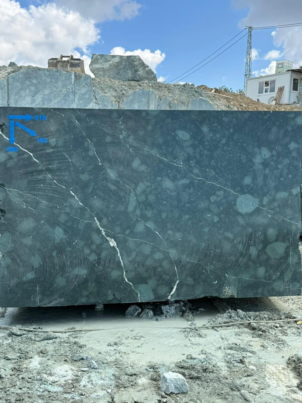 Large dark green marble slab with white veining, marked with blue measurement lines, at a construction site with dirt and rocks in the foreground and a partly cloudy sky above.