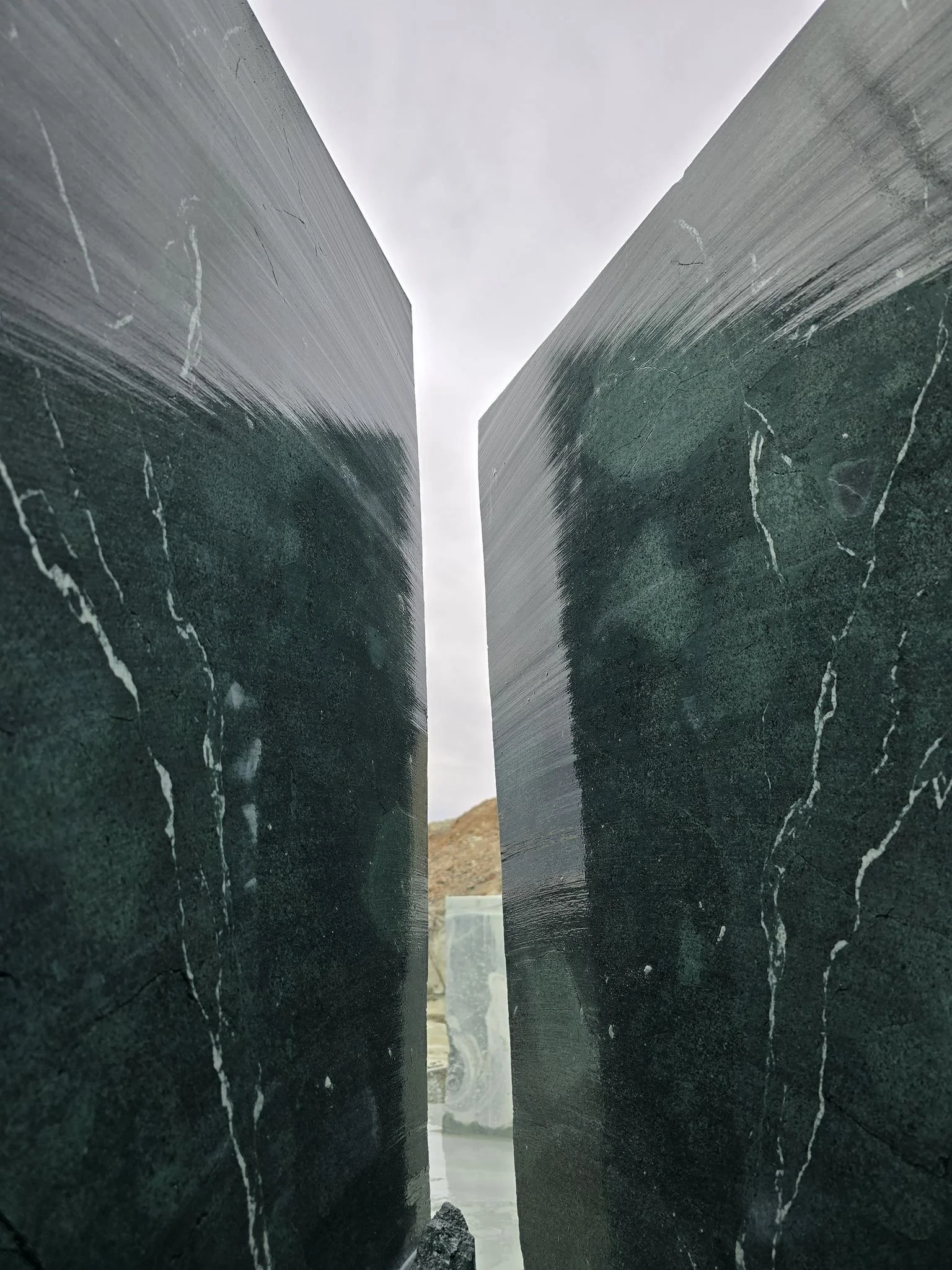 Close-up photo of two large dark green marble slabs with white veining, standing upright and separated, with a cloudy sky in the background.