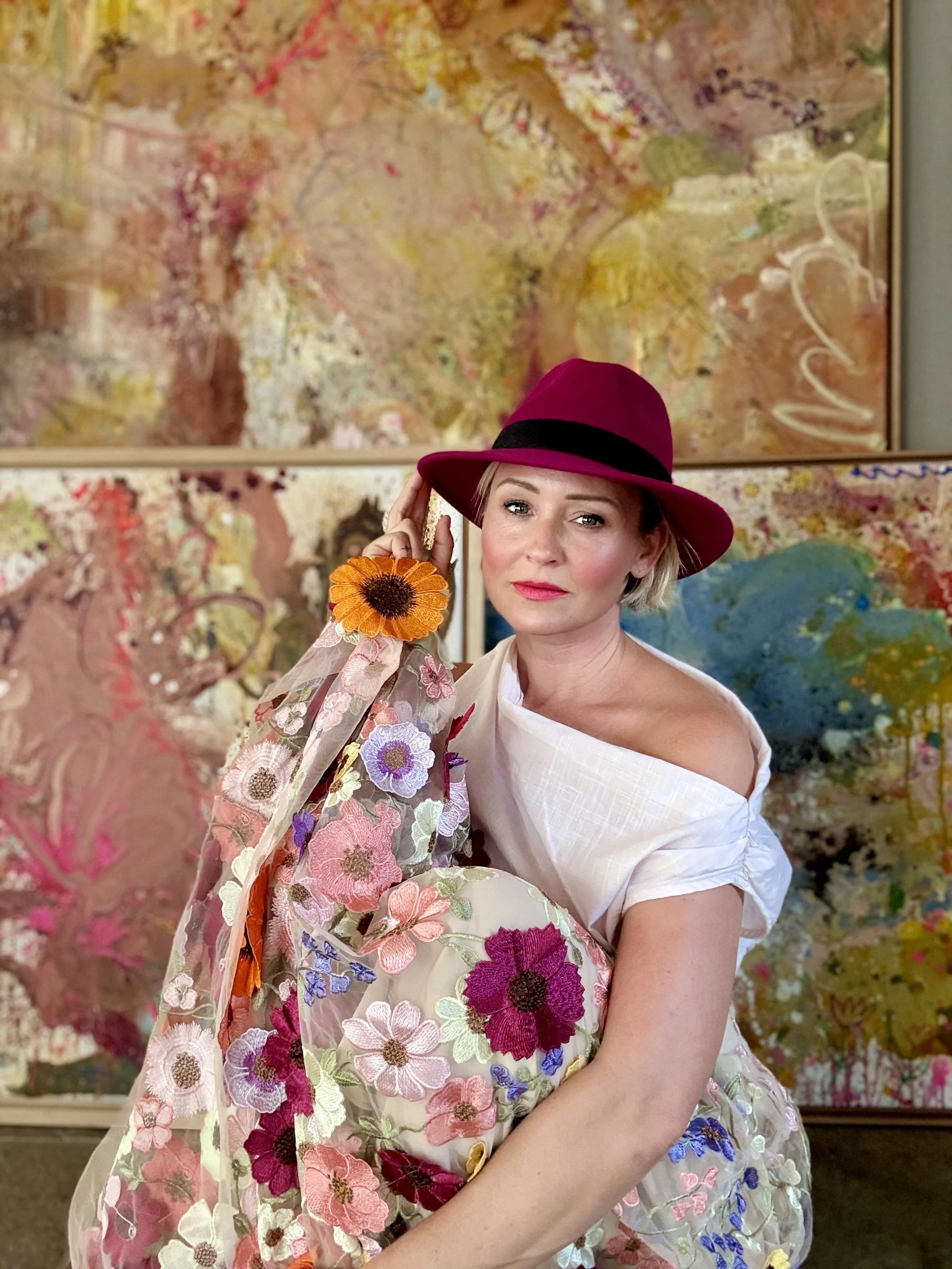Eine Frau mit kurzer blondierter Frisur, rotem Hut und rosa Lippen trägt eine weiße Kleidung mit bunten Blumenmuster. Sie steht vor einem abstrakten, farbenfrohen Kunstwerk.