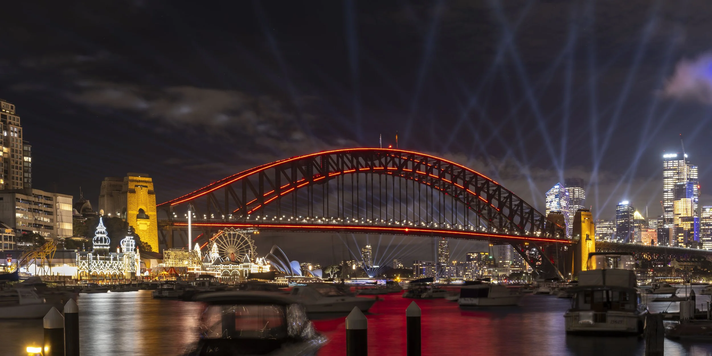 Nighttime cityscape of Sydney, Australia, featuring the illuminated Harbour Bridge with red lights, a Ferris wheel, and skyscrapers with bright lights.