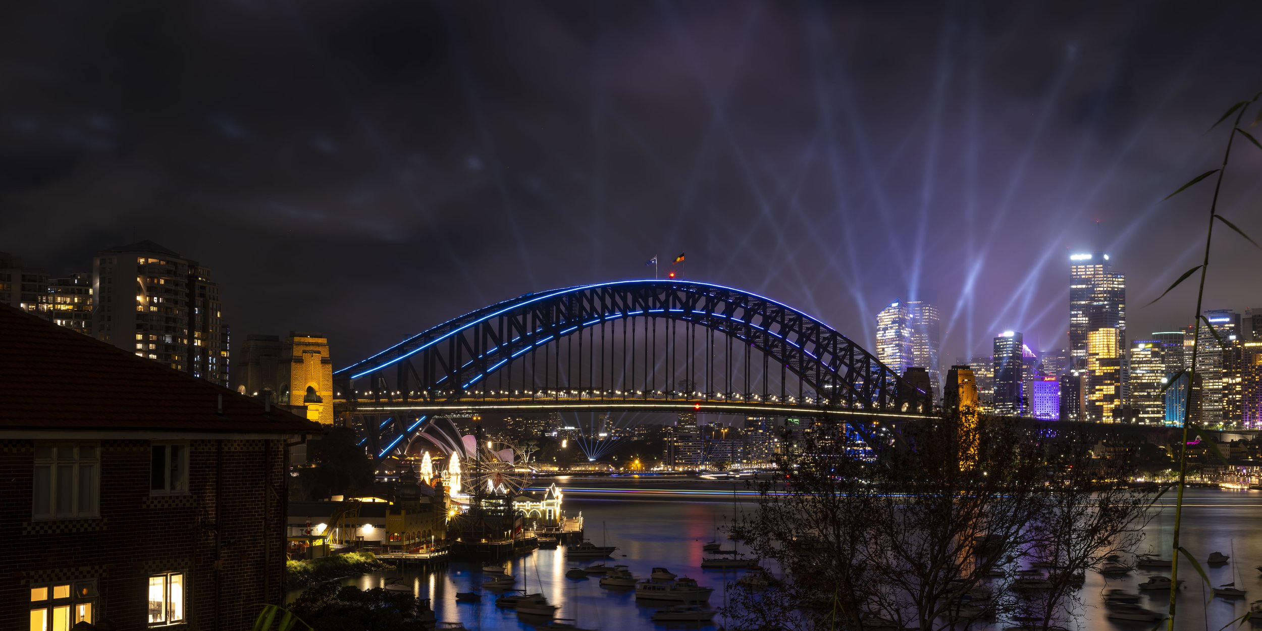 Night view of the Sydney Harbour Bridge illuminated with blue lights, city skyline with skyscrapers and colorful lights, fireworks in the sky, reflections on the water, some buildings with lit windows, boats in the harbor.