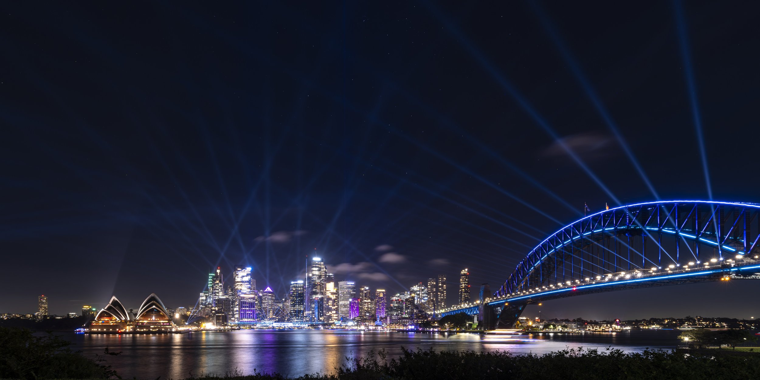 Night view of the Sydney skyline with the Sydney Opera House, Harbour Bridge, and city buildings illuminated, and light beams shooting into the sky.