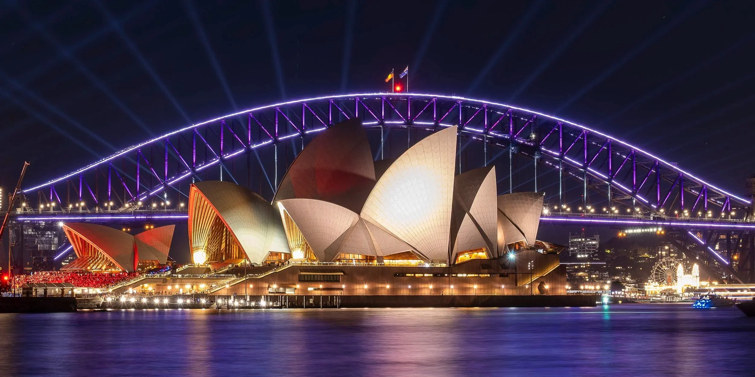 Night view of the Sydney Opera House and Sydney Harbour Bridge illuminated with colorful lights, with water in the foreground reflecting the lights.