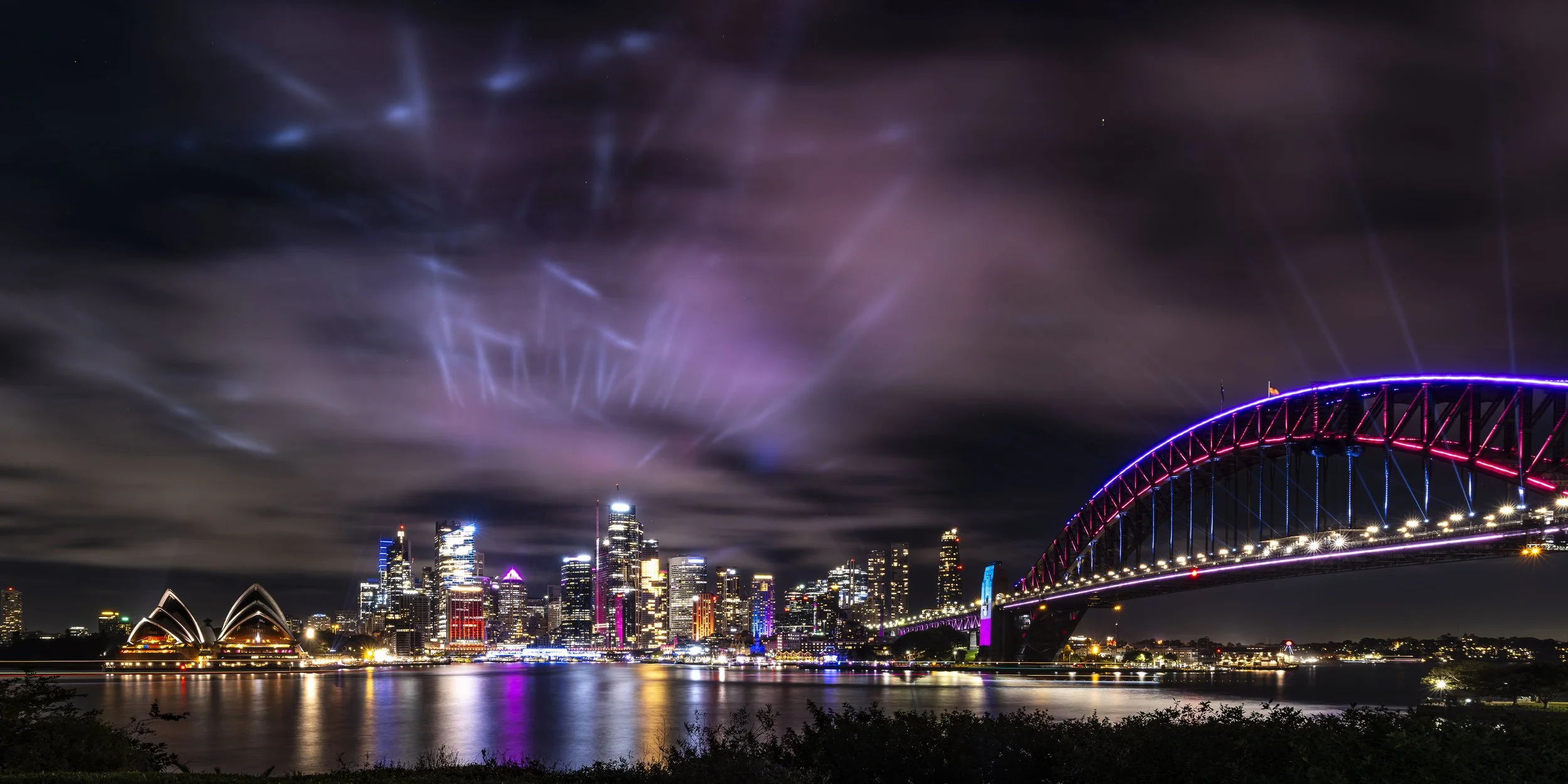 Nighttime view of a city skyline with buildings lit up, a bridge with colorful lights, and fireworks in the sky reflected on the water.