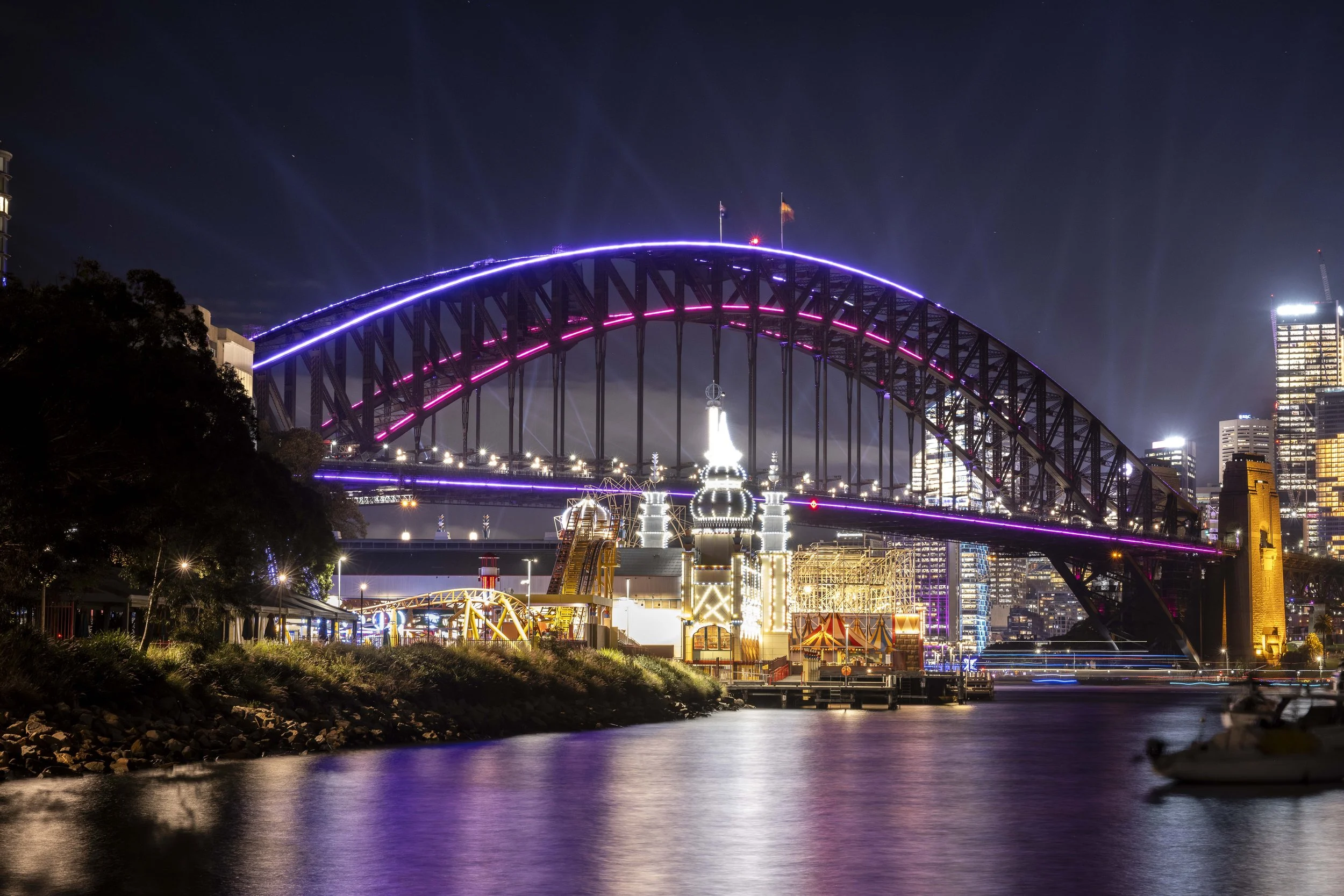 Night view of the Sydney Harbour Bridge illuminated with purple and pink lights, reflecting on the water below, with city skyline and buildings lit up in the background.