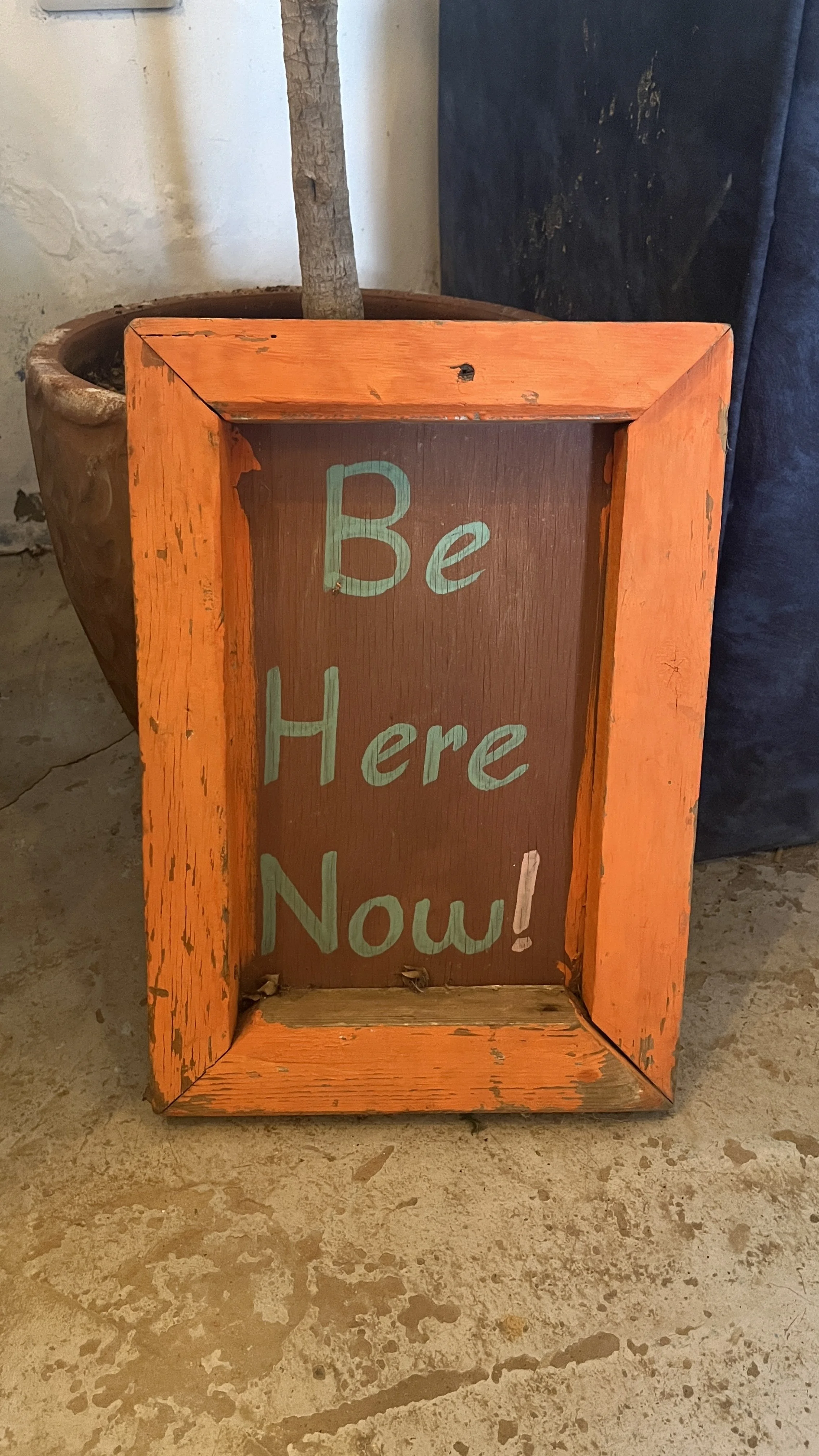 A wooden sign with an orange painted frame and a brown interior that reads 'Be Here Now!' in green text, leaning against a wall with a potted plant in the background.