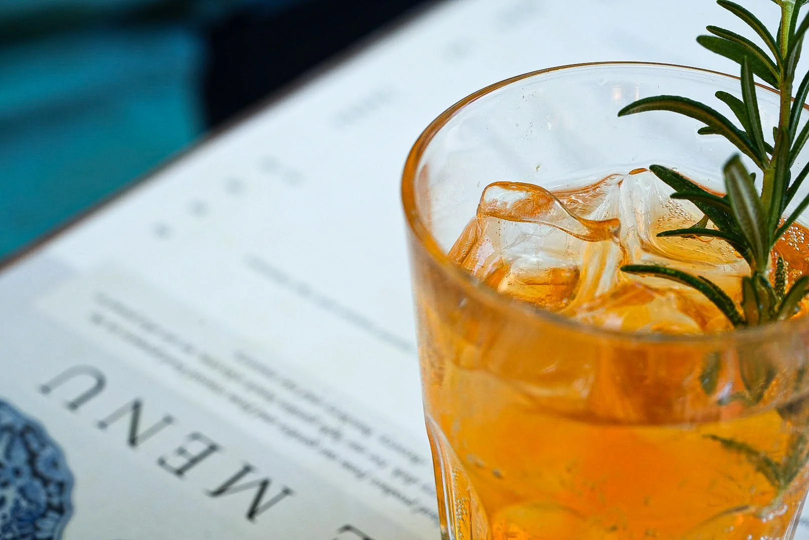 Produktfotografie von einem Glas mit Orangenlimonade, Eiswürfeln und einem Zweig Rosmarin.