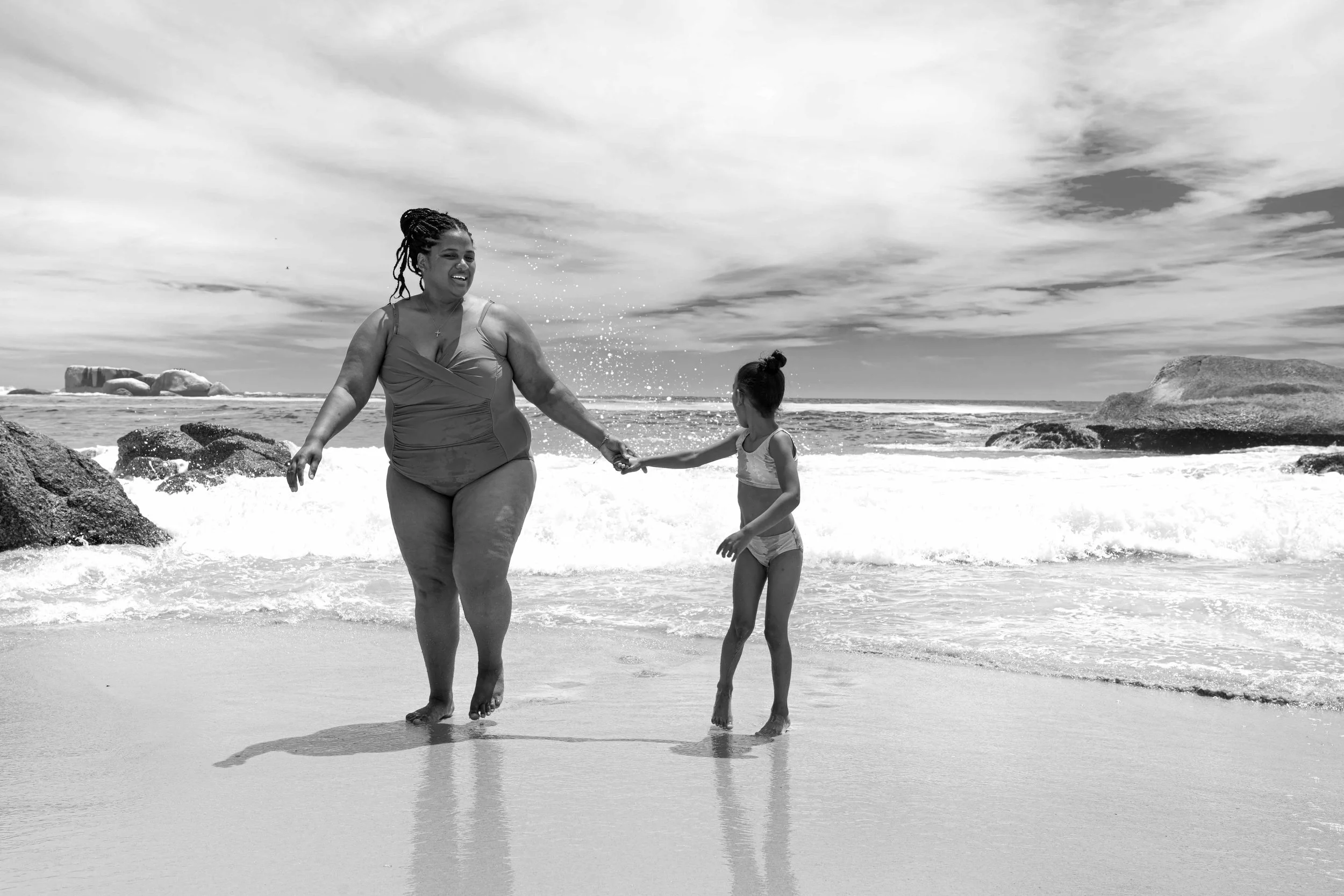 Streetphotograohy von einer Frau und einem Kind am Strand, halten sich an den Händen, das Wasser spritzt beim Laufen, im Hintergrund Himmel mit Wolken, Meer und Felsen, schwarz-weiße Fotografie.