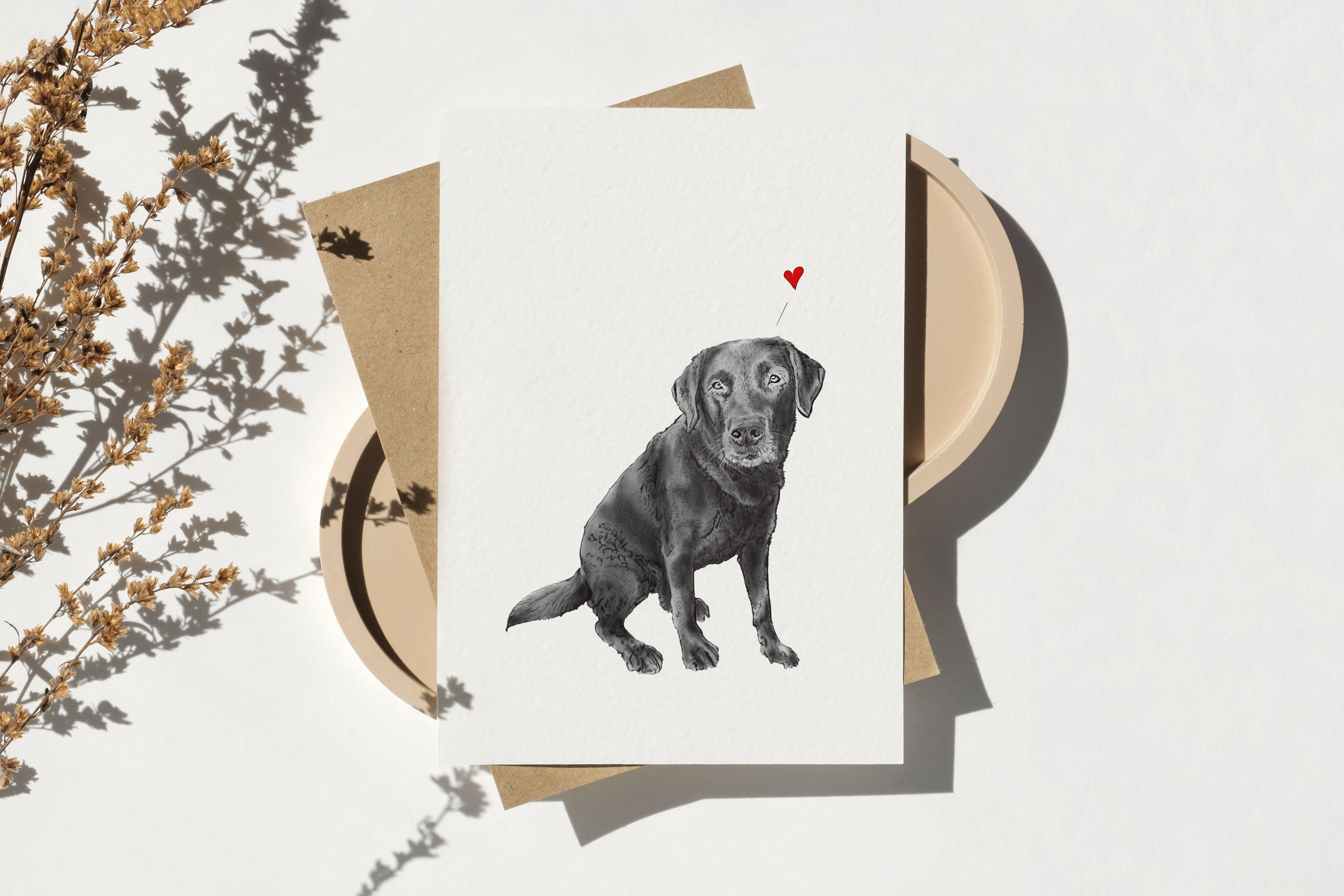 Schwarz-weiß Zeichnung eines Hundes mit kleinem roten Herz darüber, auf einem weißen Blatt Papier liegend, mit getönten Papierstücken und trockenen Blumen daneben.