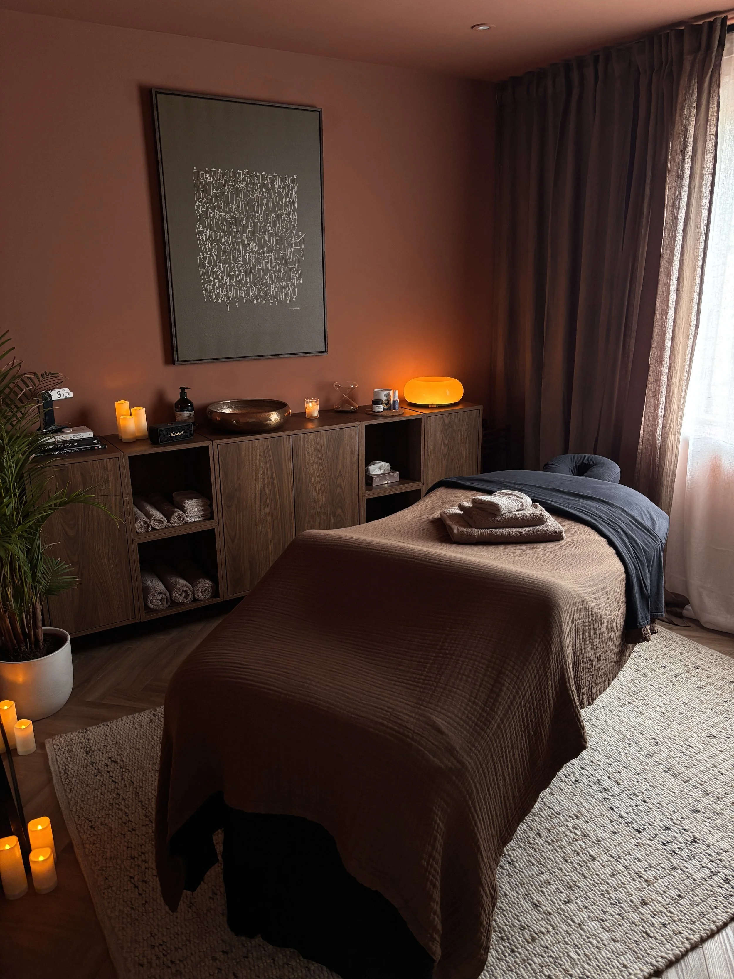 A cozy massage room with a massage table covered in a brown blanket, folded towels on top, surrounded by warm lighting, candles, a wooden cabinet with decorative items, candles, and a glowing lamp, with dark curtains and a piece of modern wall art.