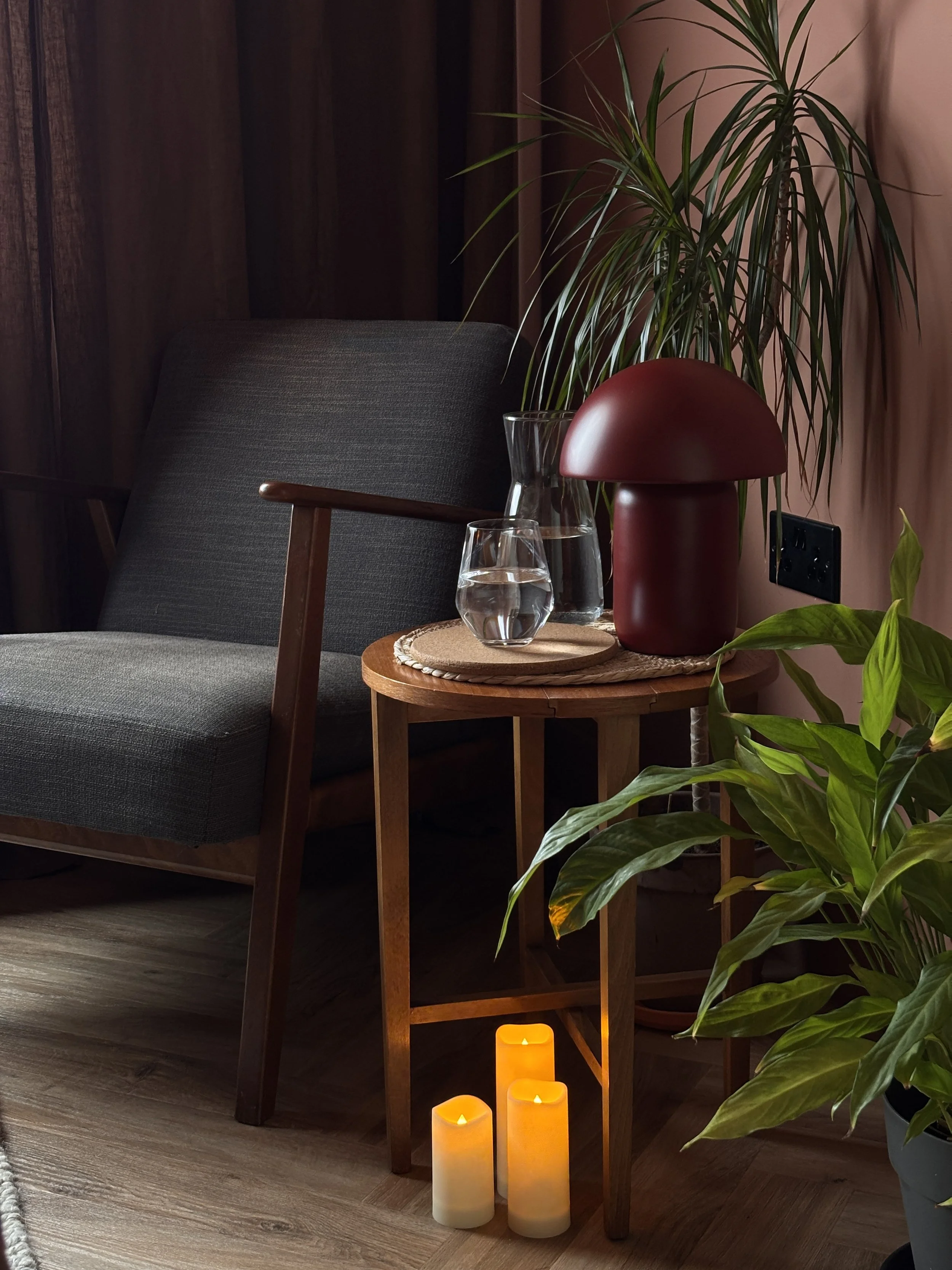 Cozy corner with a dark gray upholstered armchair, a small wooden side table, a large potted plant, a red mushroom-shaped table lamp, glass vase with water, and yellow LED candles on the floor, all in a warmly lit room.