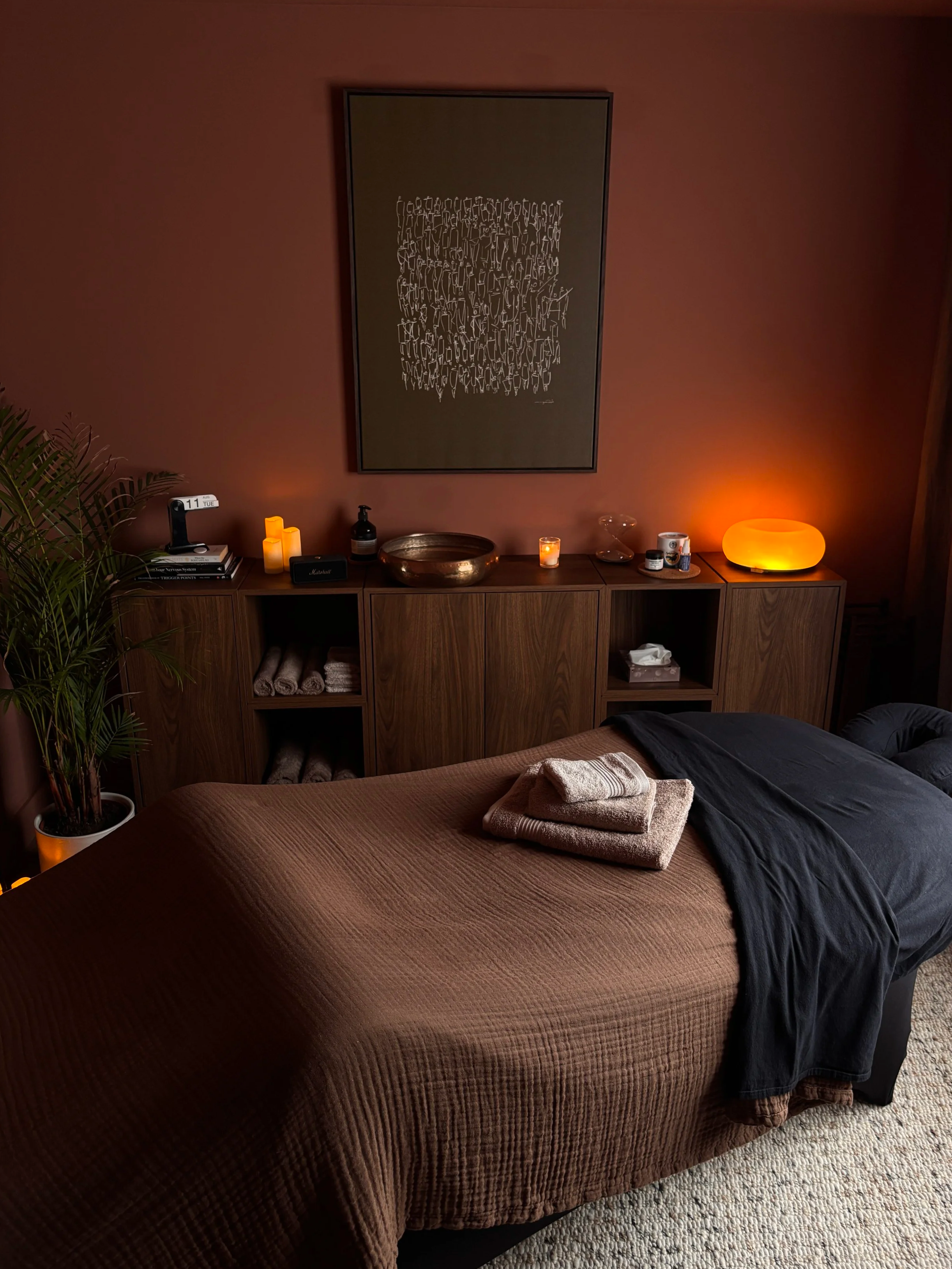 A bedroom with a bed covered in a brown quilt and a black throw. Towels are folded on the bed. There is a wooden sideboard with various items, including candles, a plant, a lamp, and personal care products. A large framed abstract art piece hangs above the sideboard, and a textured area rug is partially visible on the floor.