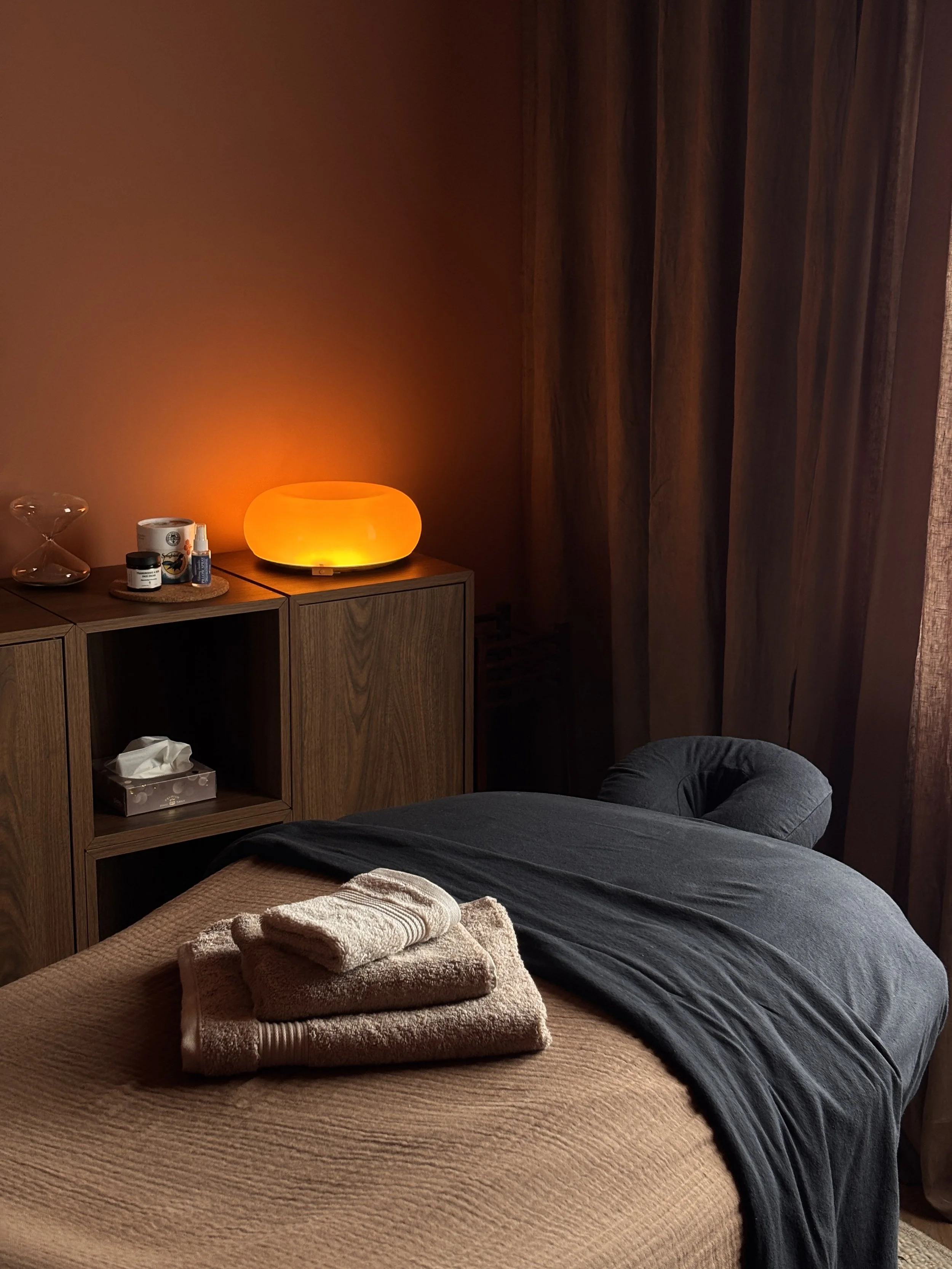 A bedroom with a bed covered in a beige textured blanket, with folded towels for a guest. A dark grey or black blanket is draped over part of the bed. To the left of the bed is a wooden cabinet with a glowing orange lamp on top, along with skincare and beauty products. The background features a dark wooden panel or wall with curtains.