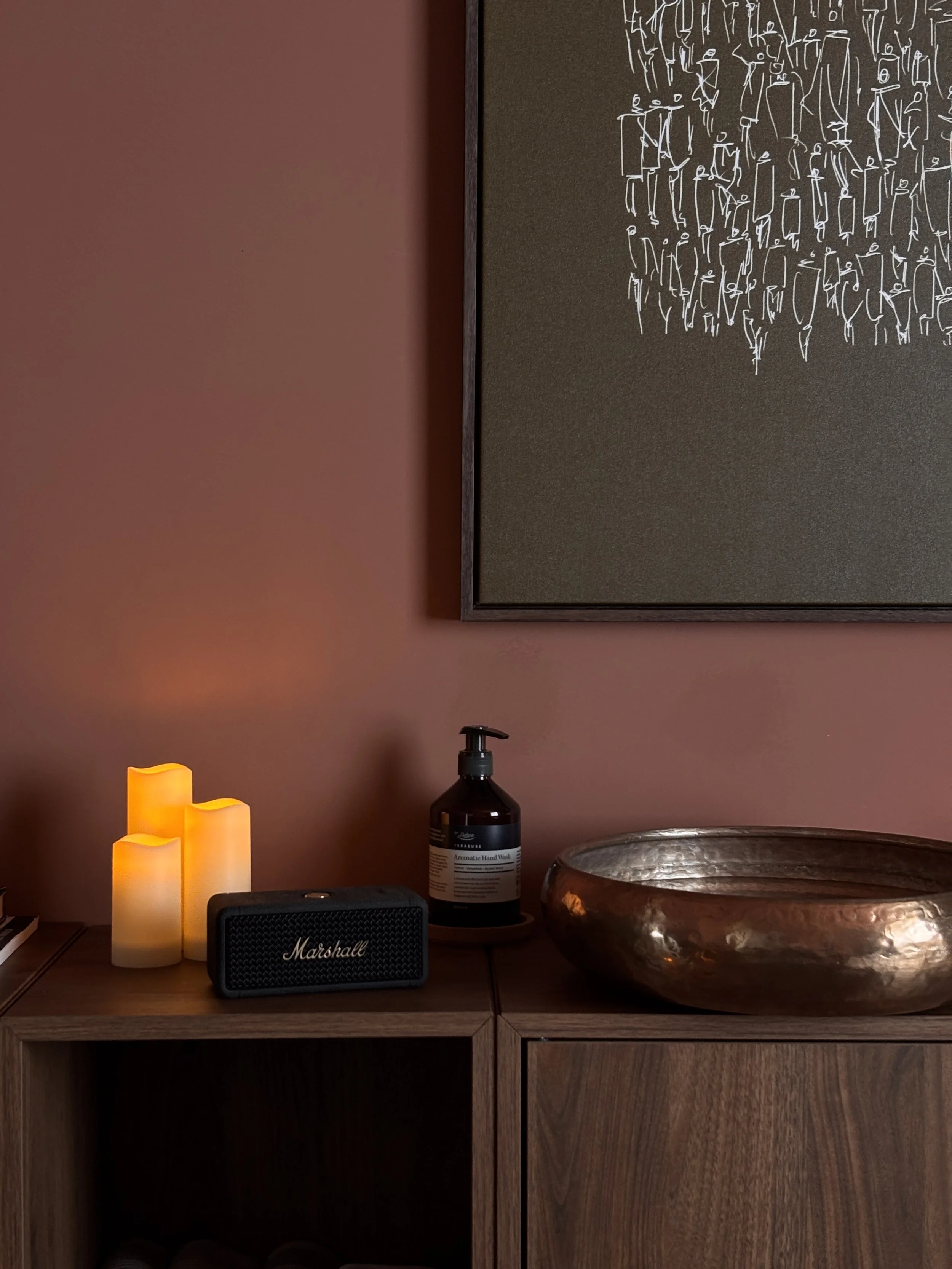 A wooden cabinet with decorative items on top, including three yellow battery-operated candles, a black speaker with 'Marshall' written on it, a bottle of aromatic hand wash, a large metallic bowl, and a framed chalkboard with white abstract line art on a dark background hanging on a pink wall.