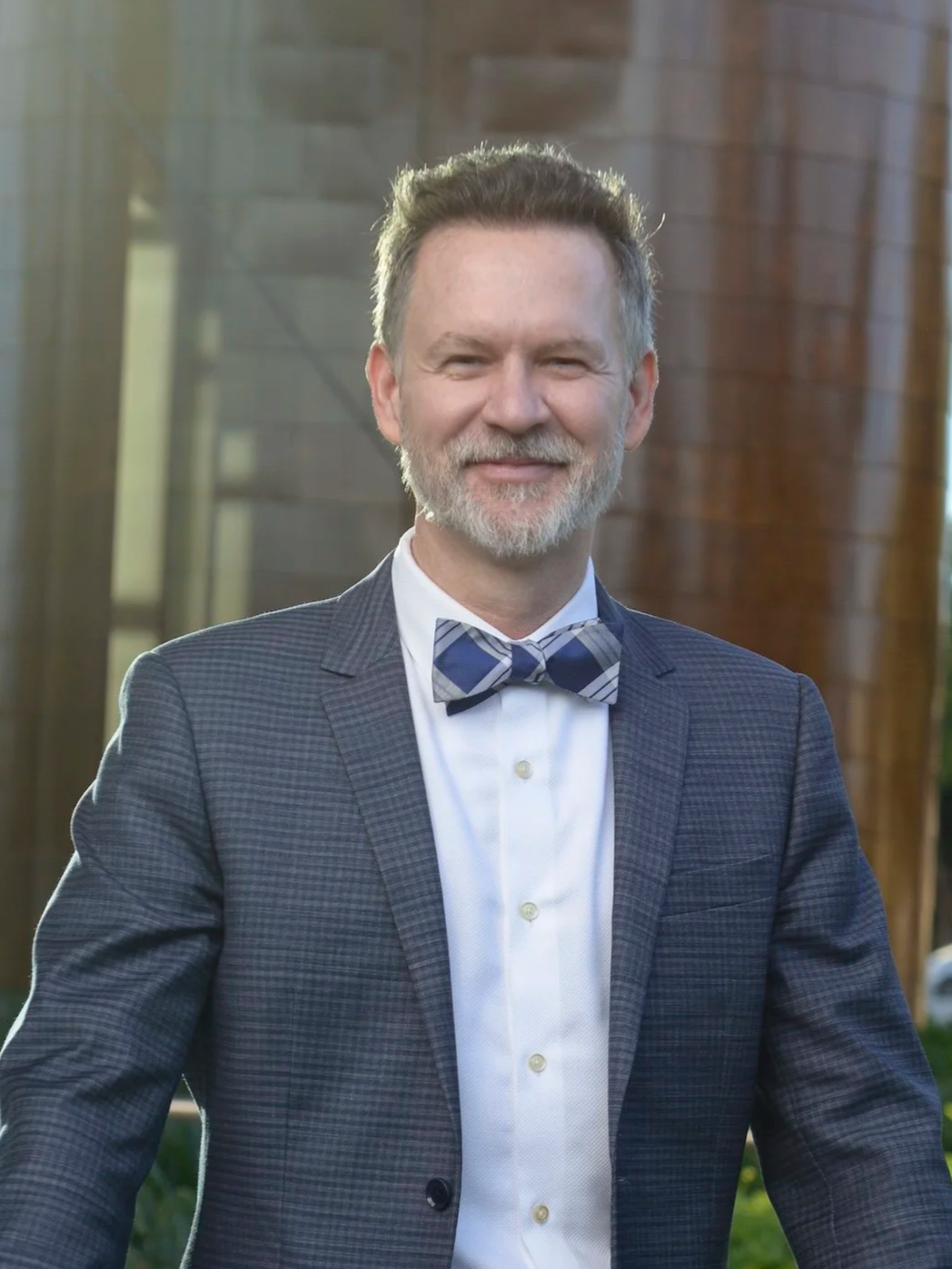 A smiling man with gray hair and beard, dressed in a checkered suit jacket, white dress shirt, and a plaid bow tie, standing outdoors with modern buildings in the background.
