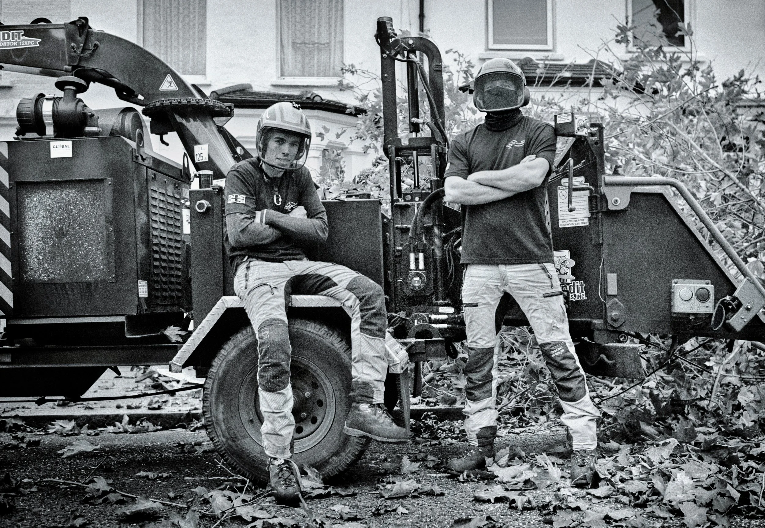 Two workers in helmets and work clothes pose in front of a wood chipper machine on a leaf-covered street.
