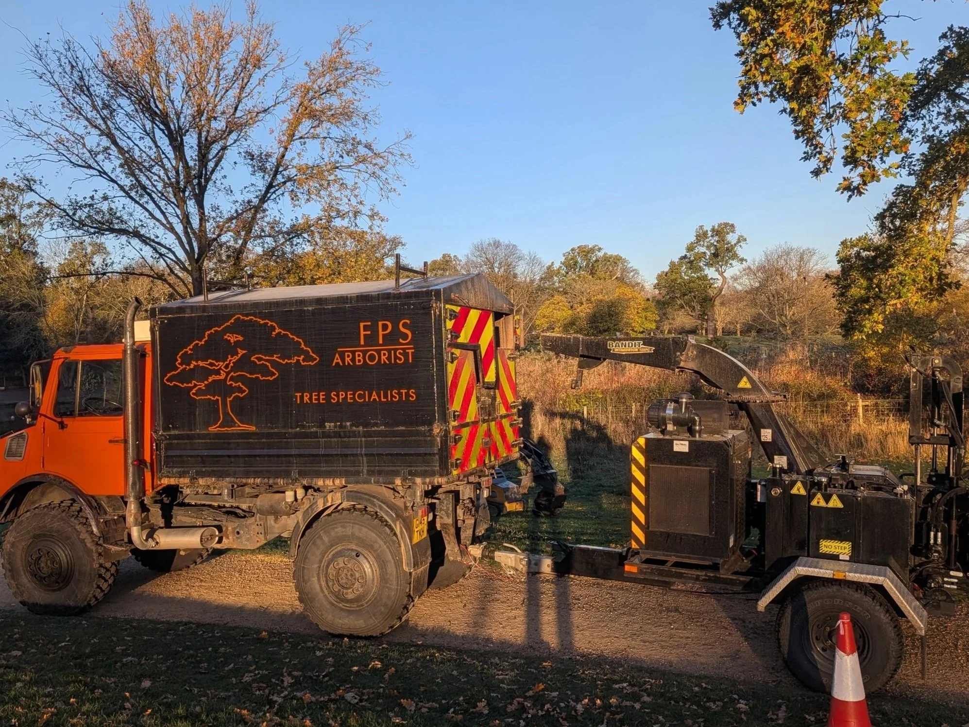 A tree trimming truck with the logo 'FPS Arborist Tree Specialists' parked on a dirt path, with tree pruning equipment attached, during fall with leafless trees and a clear blue sky in the background.