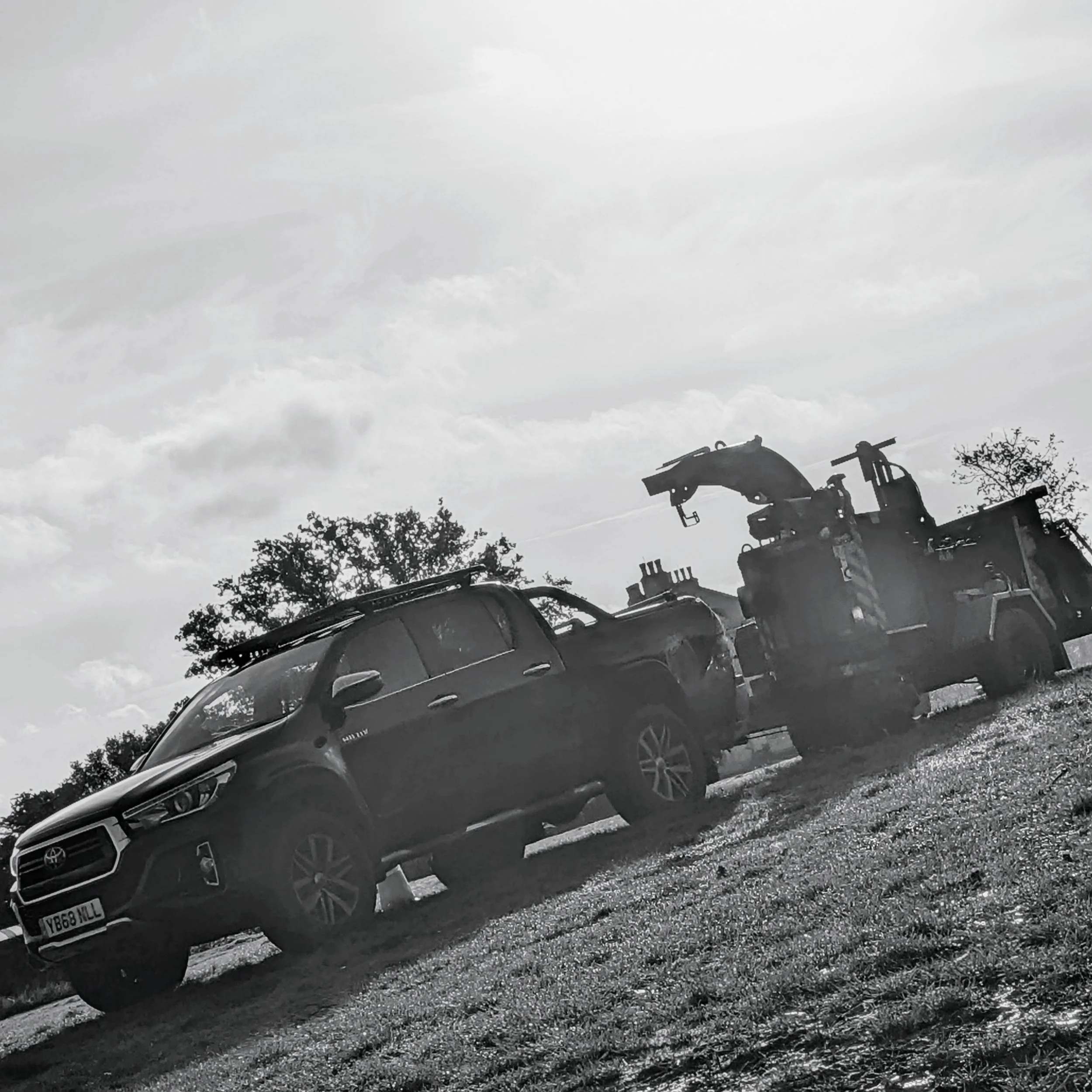 Black-and-white photo of a parked SUV and a tow truck with a crane, with trees and clouds in the background.