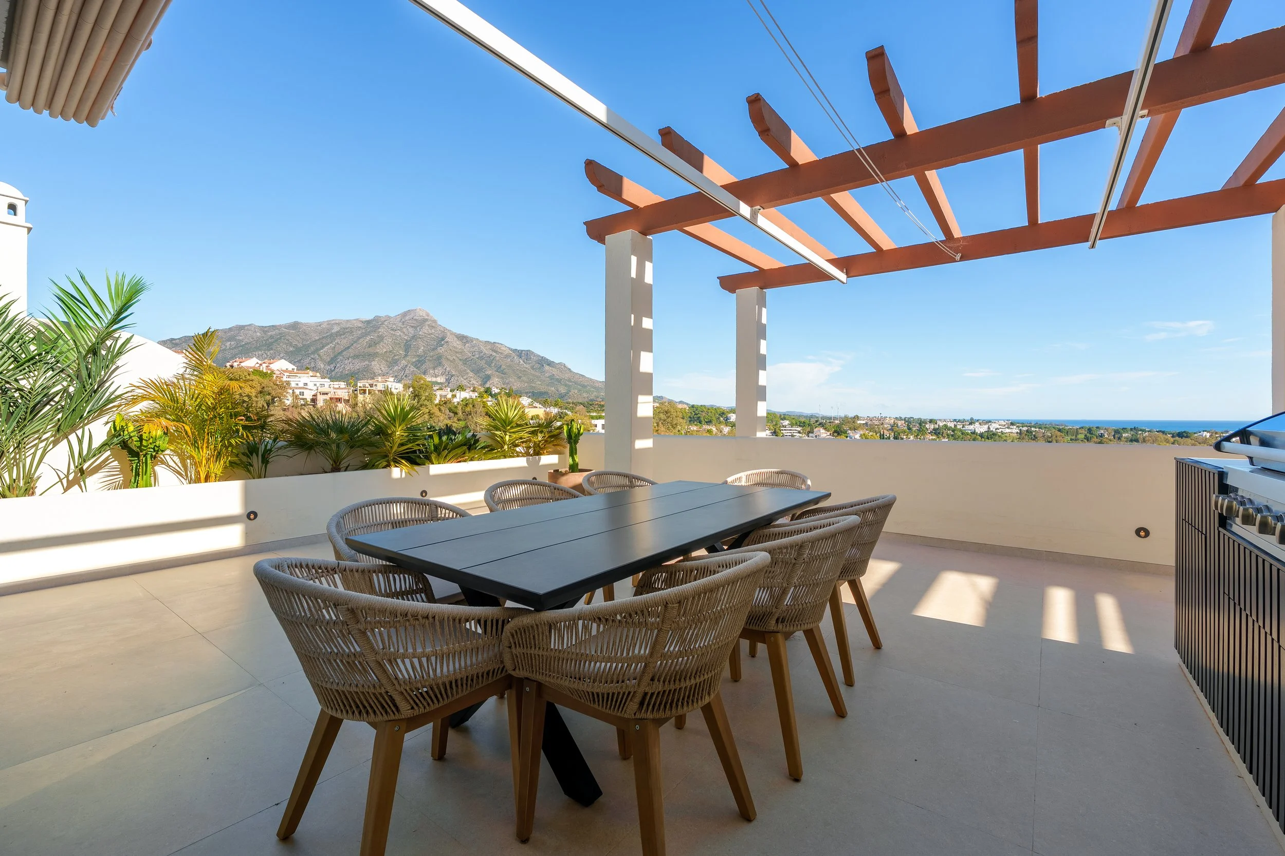Covered terrace dining area with a dark wood table and wicker chairs, perfect for al-fresco meals by the outdoor kitchen.