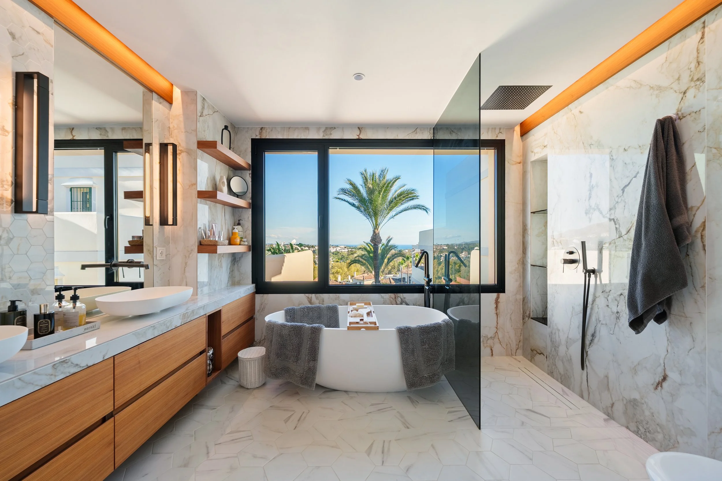 Luxurious bathroom featuring a freestanding oval tub, double vanity with wooden accents, and a large window framing tropical views.
