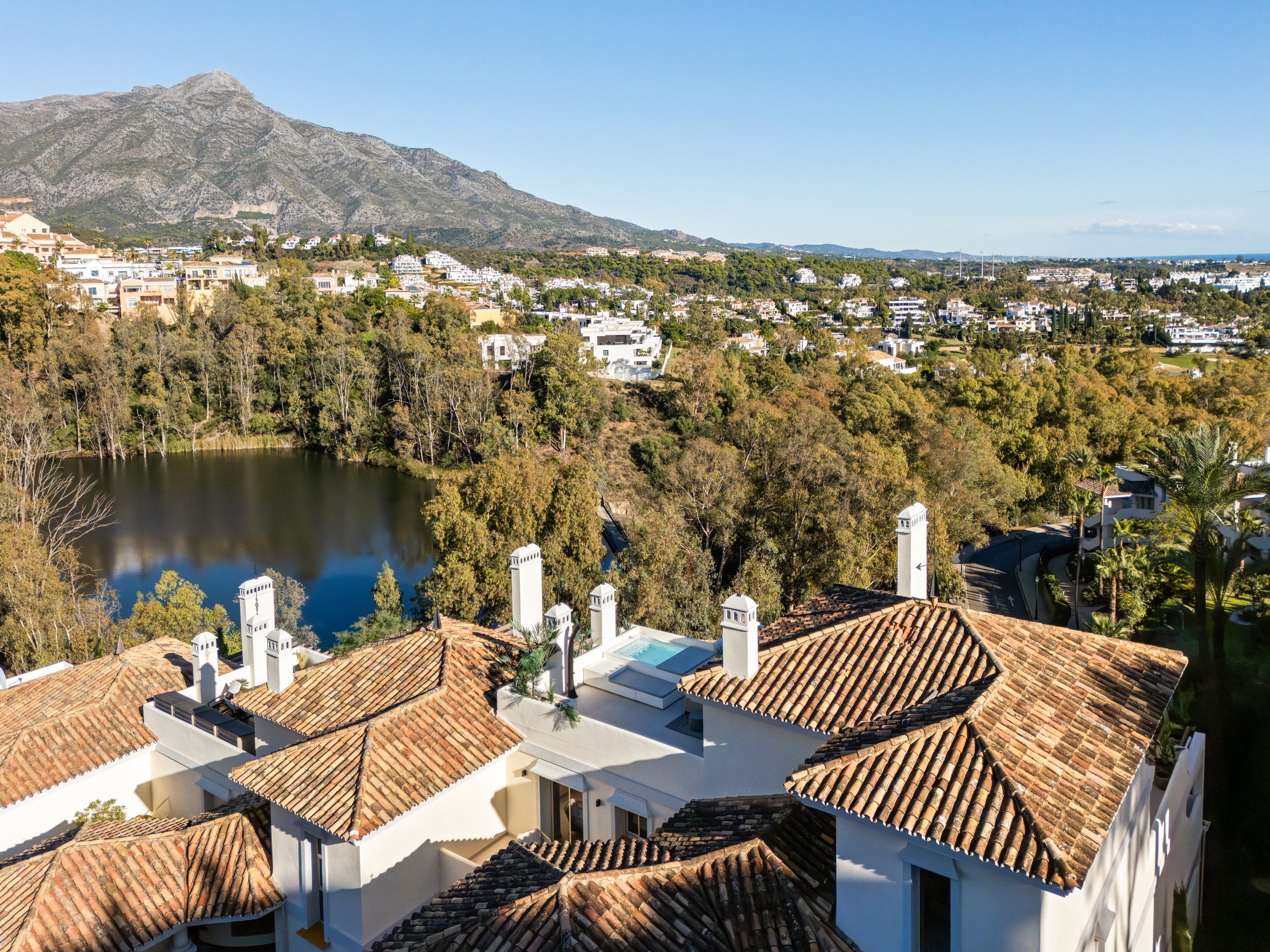 Elevated panoramic view from the penthouse overlooking the lush greenery of Nueva Andalucía, a peaceful lake, and the surrounding mountains.