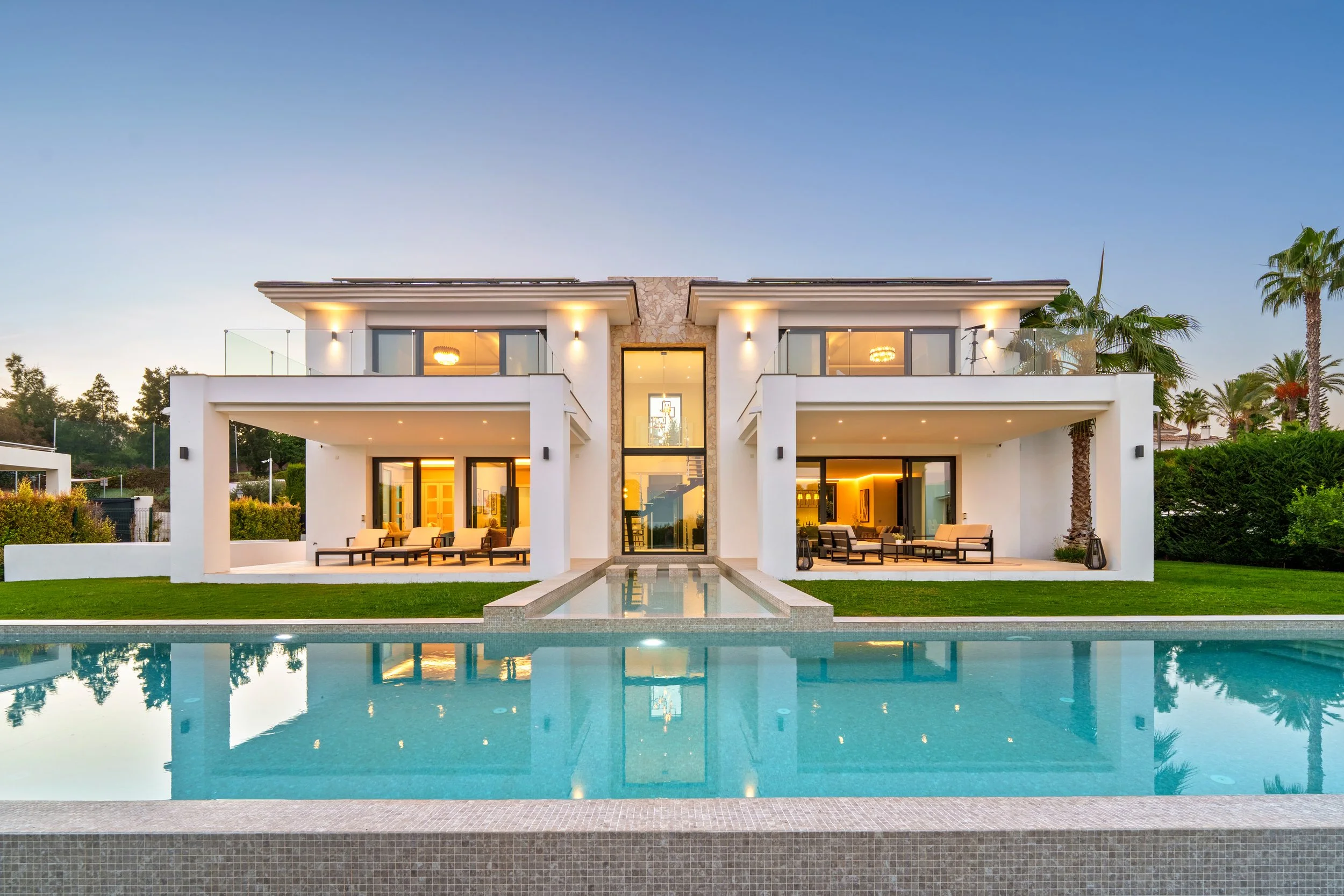 Twilight view of a modern white villa in La Quinta with an illuminated infinity pool and symmetrical architecture.