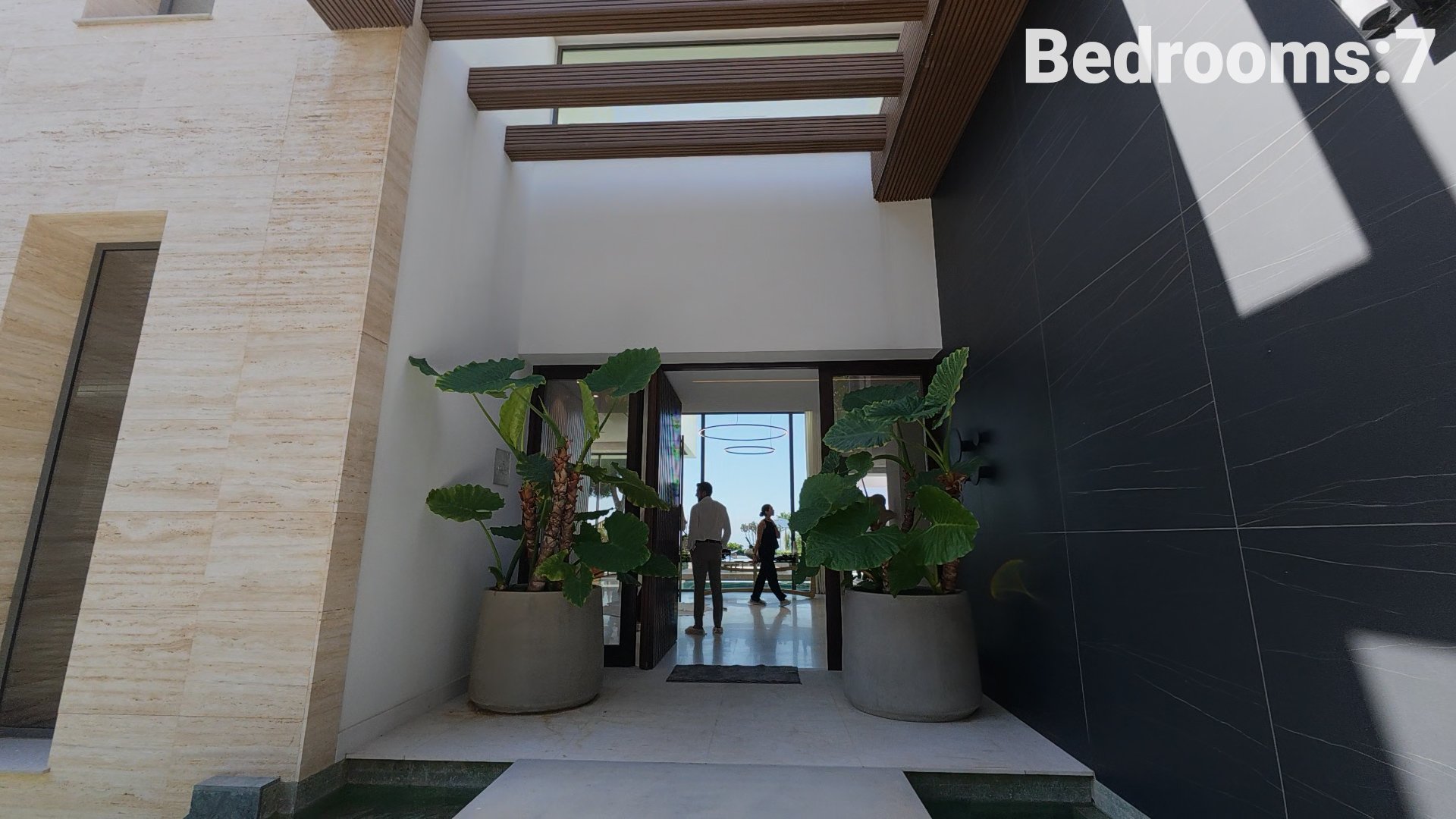 Walk-through angle during the property tour focusing on the contemporary glass main entrance framed by modern stone pillars and abundant greenery.