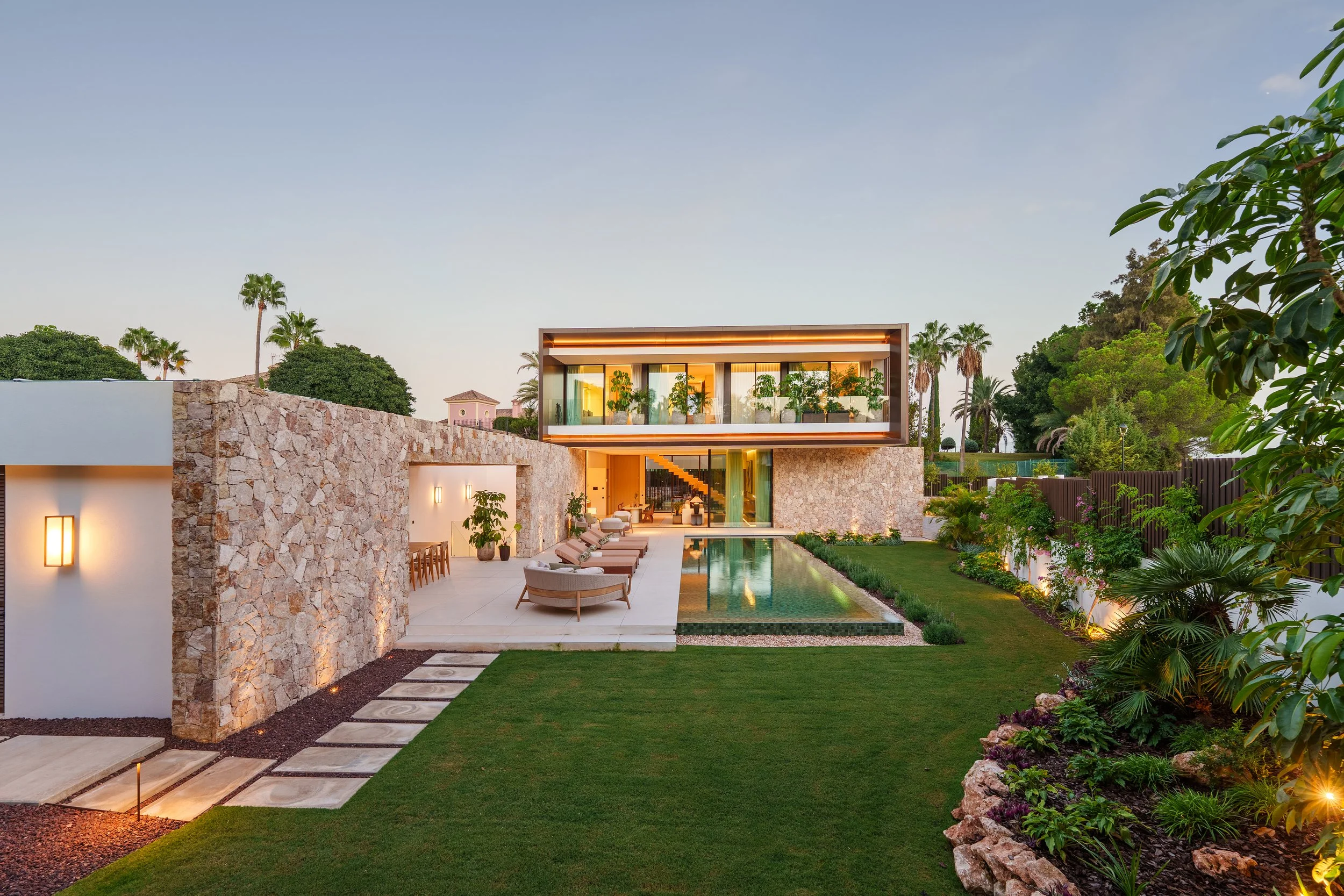 A luxury contemporary villa at dusk featuring a stone facade, an illuminated infinity pool, and a manicured green lawn.