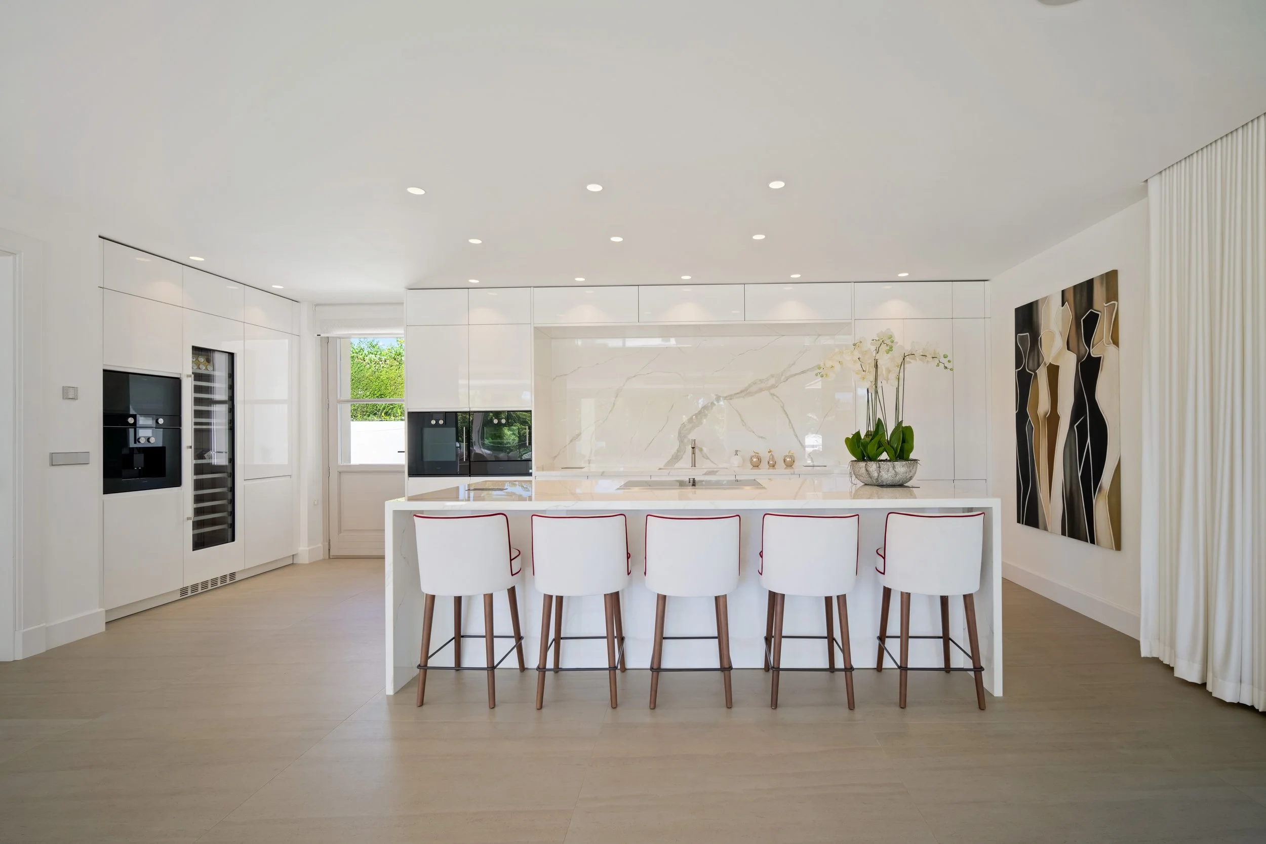 Modern minimalist white kitchen featuring a large central island with seating for five, integrated appliances, and clean architectural lines.
