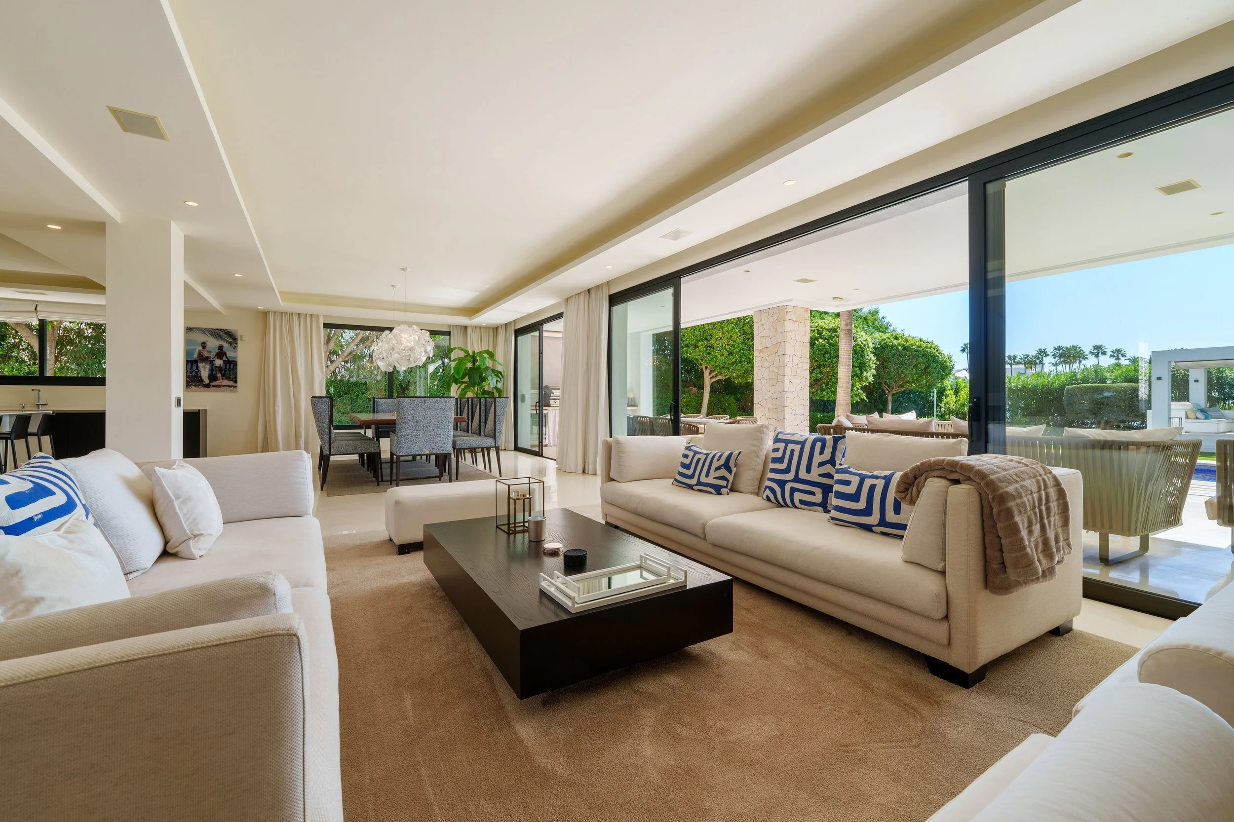 Open-plan luxury living area with floor-to-ceiling glass doors, neutral contemporary furniture, and seamless indoor-outdoor flow to the pool terrace.