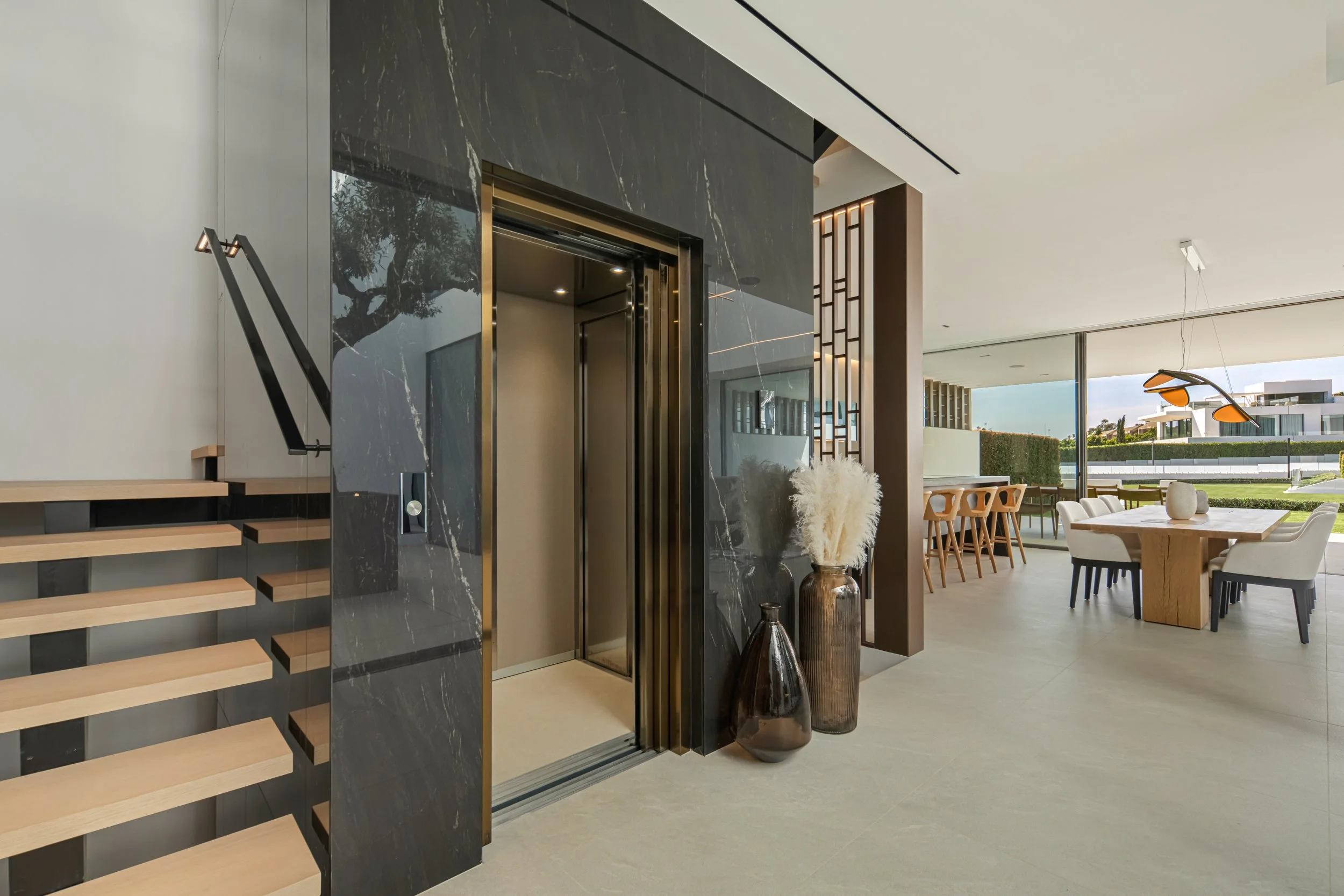 Modern interior hallway featuring a private elevator clad in dark stone and a floating wooden staircase.