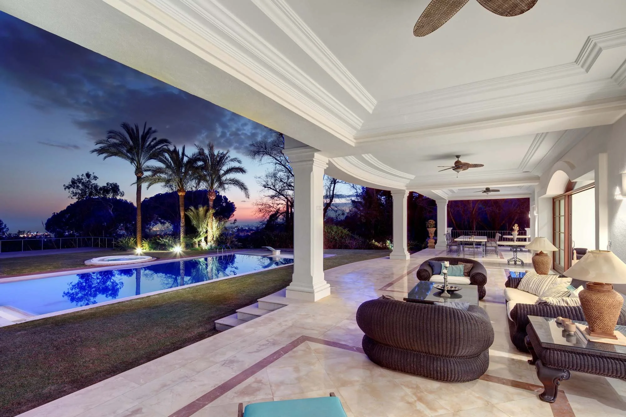 Luxury covered terrace lounge with outdoor fans, marble tiling, and heated pool views at dusk.