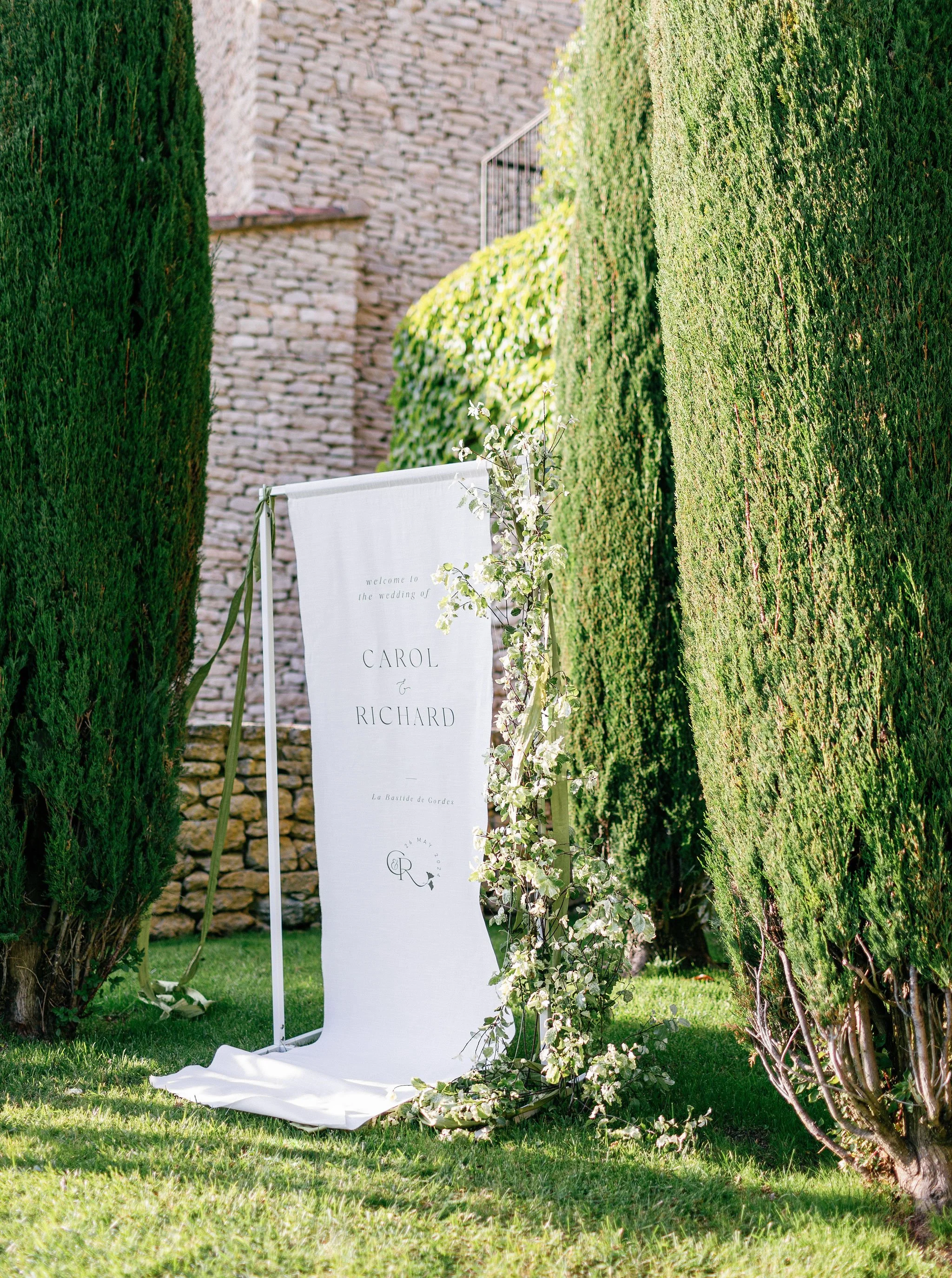 Wedding welcome sign with floral decorations, placed between tall green hedge plants on a grassy area.