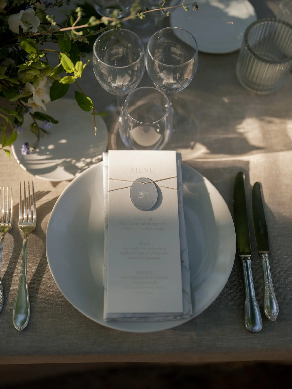 Elegant table setting with a white plate, vintage silver cutlery, three wine glasses, a menu, and a small bouquet of flowers in a vase.