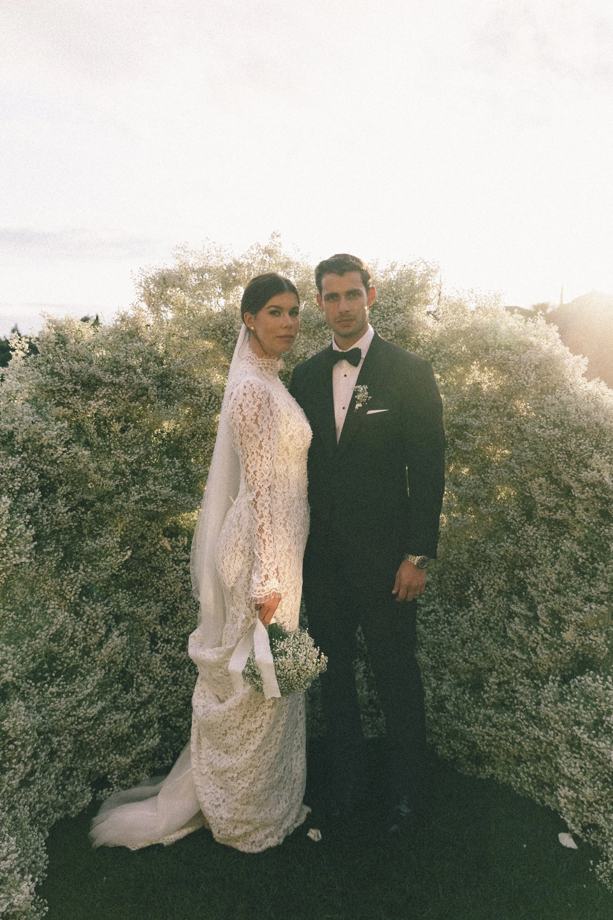 A bride and groom standing outdoors surrounded by white flowers, during sunset.