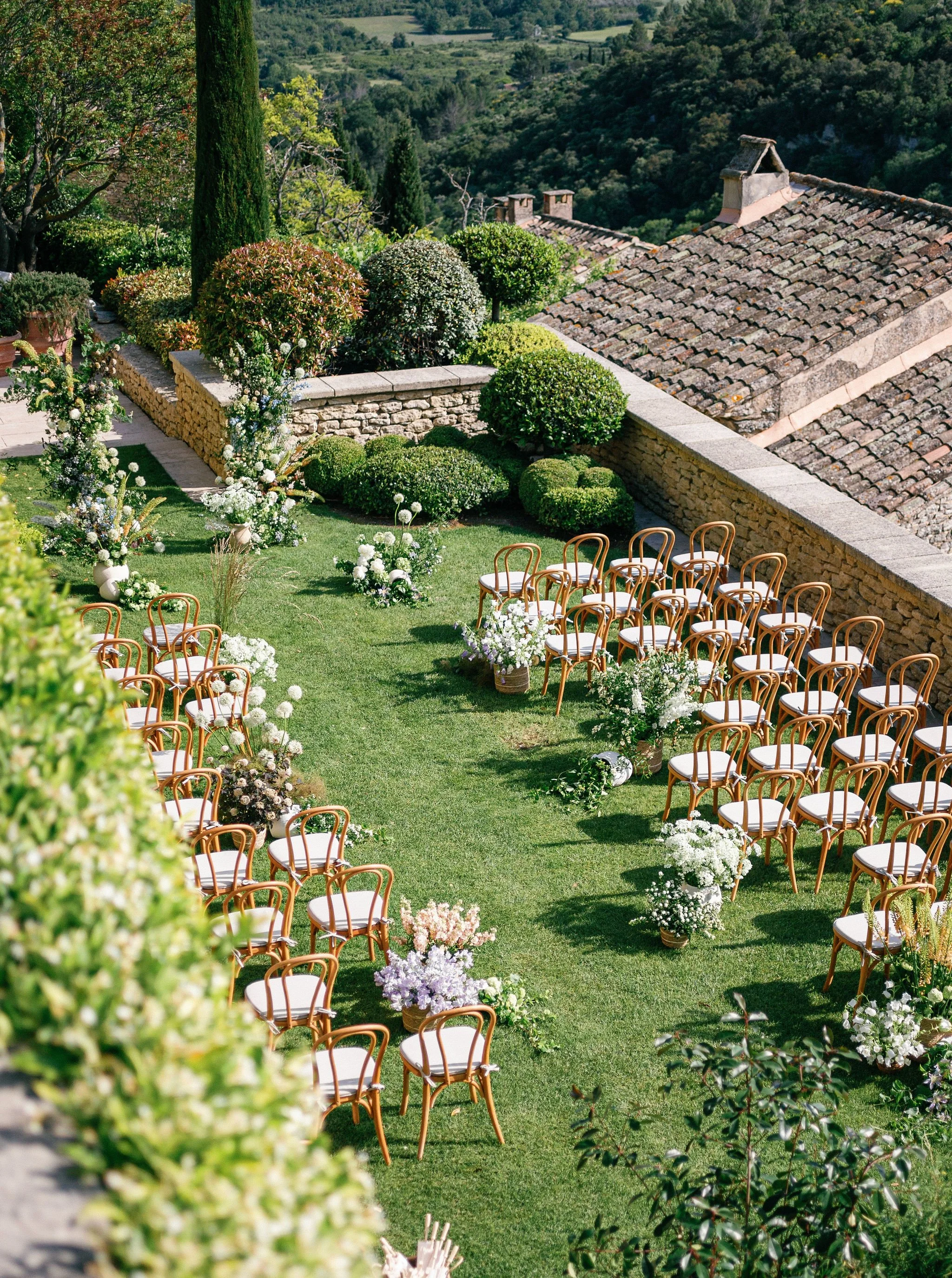 Gordes outdoor wedding ceremony setup with chairs arranged in rows on a grassy lawn, decorated with flowers and surrounded by greenery and trees, with a scenic view of hills of Gordes, Provence.