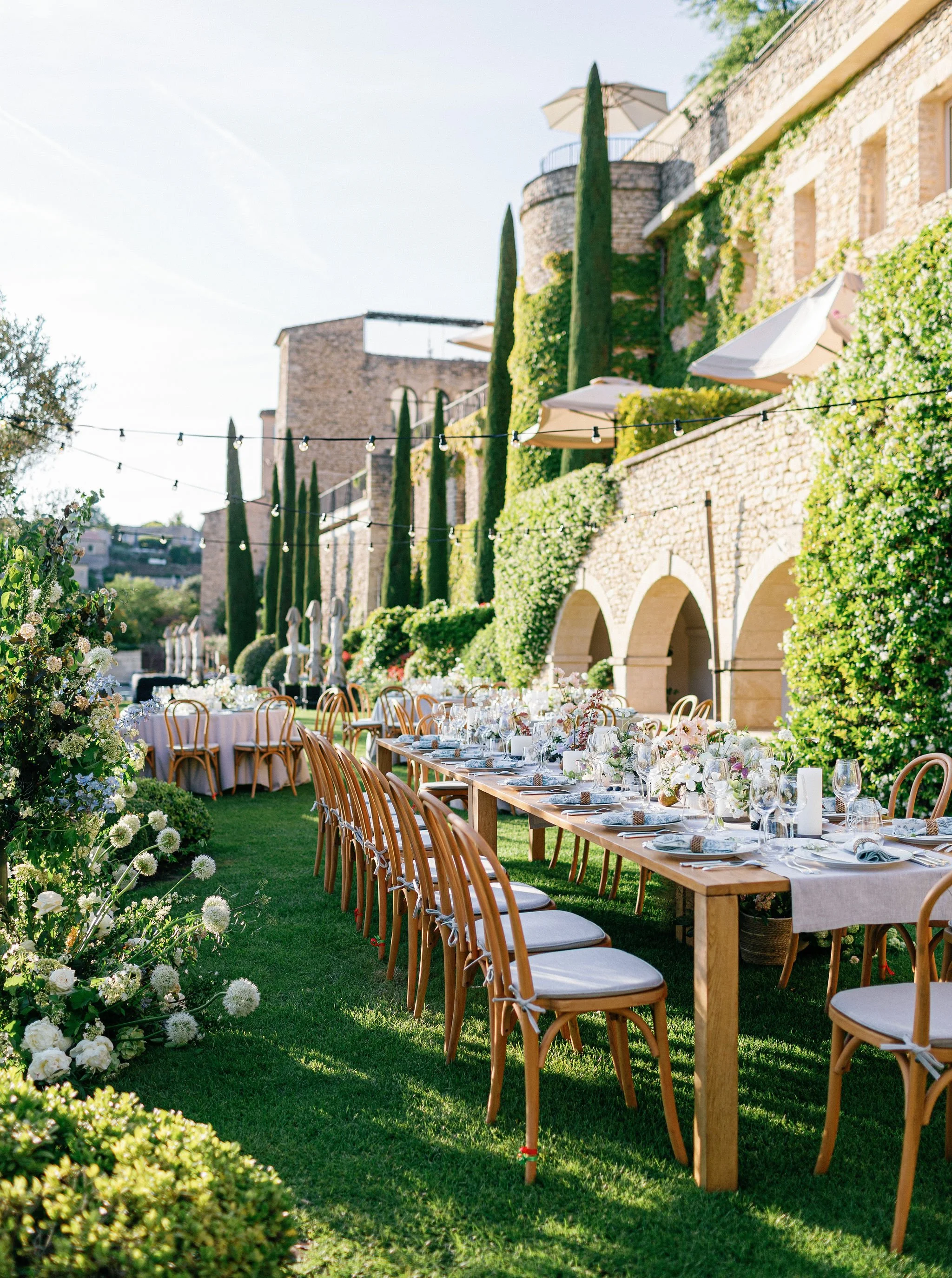 Outdoor event setup with long wooden tables decorated with white flowers and dinnerware, surrounded by wooden chairs with white cushions, lush green grass, and a stone building with arches and tall cypress trees in the background.