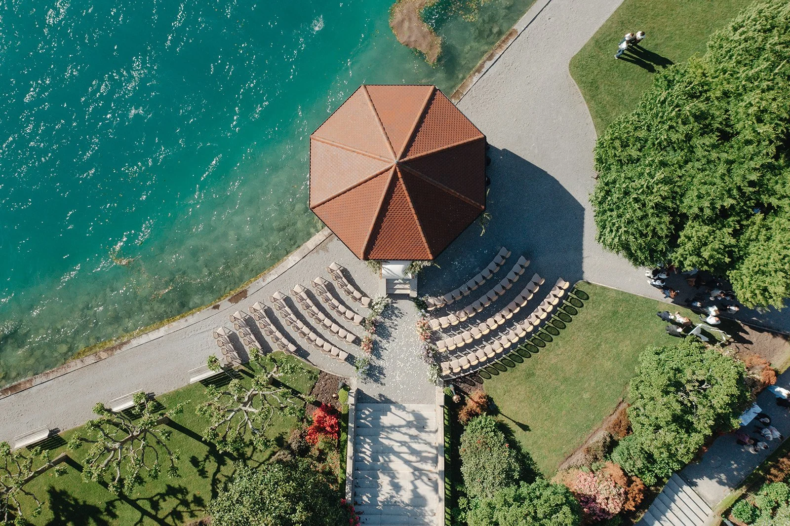 An aerial view of a gazebo by the Annecy lake, with chairs arranged in a semi-circle around it for the wedding ceremony at Palace de Menthon, green trees, and a group of people walking along a pathway.