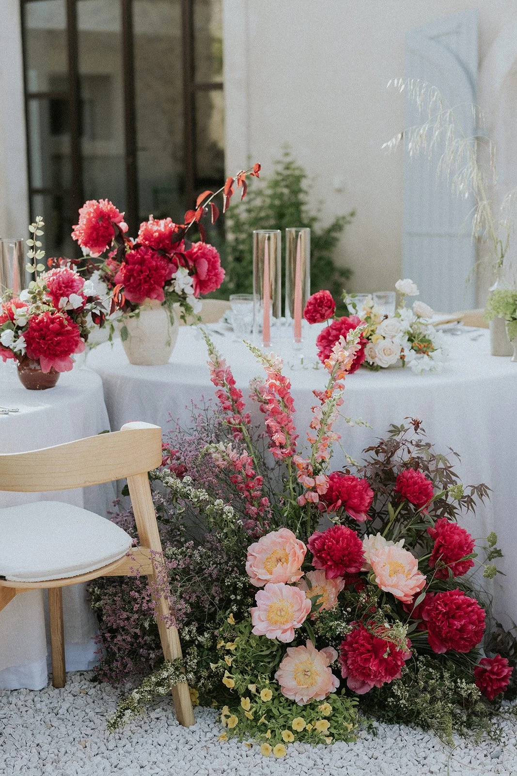 A beautifully decorated table at an outdoor event with pink and white floral arrangements, including peonies and other flowers, complemented by tall glass candle holders, on a white tablecloth surrounded by a light wooden chair.