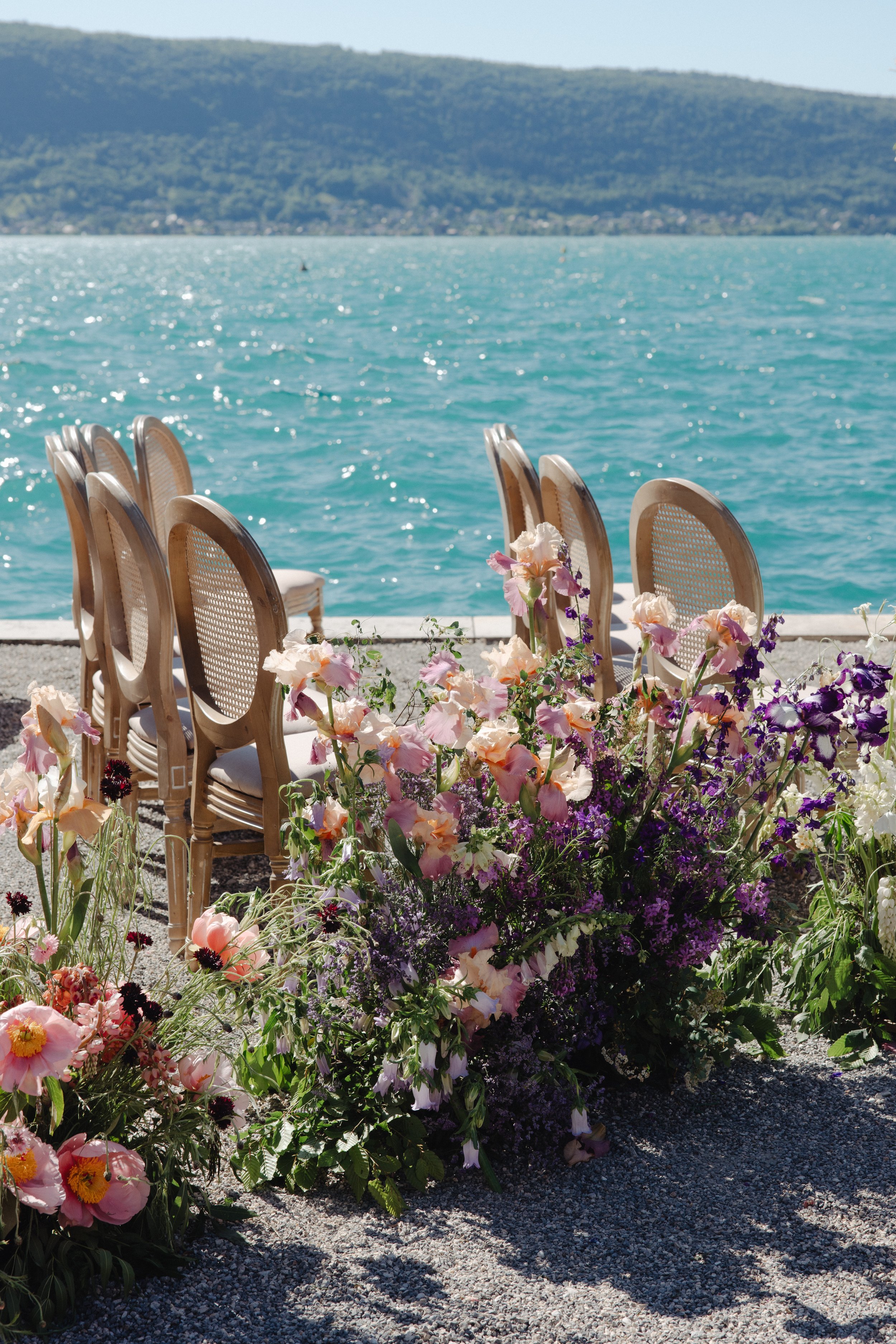 Ceremony chairs and a floral arrangement set on a gravel surface near the Annecy lake with mountains in the background.