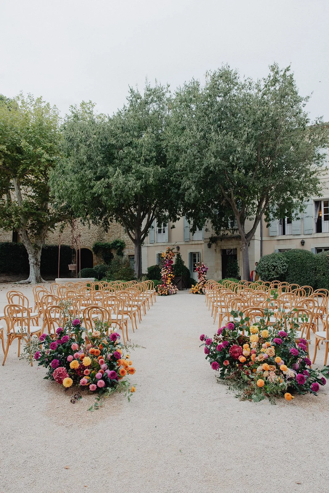 Outdoor wedding ceremony setup with rows of wooden chairs, floral arrangements on the ground, and a floral arch at the altar, surrounded by trees and a building in the background.